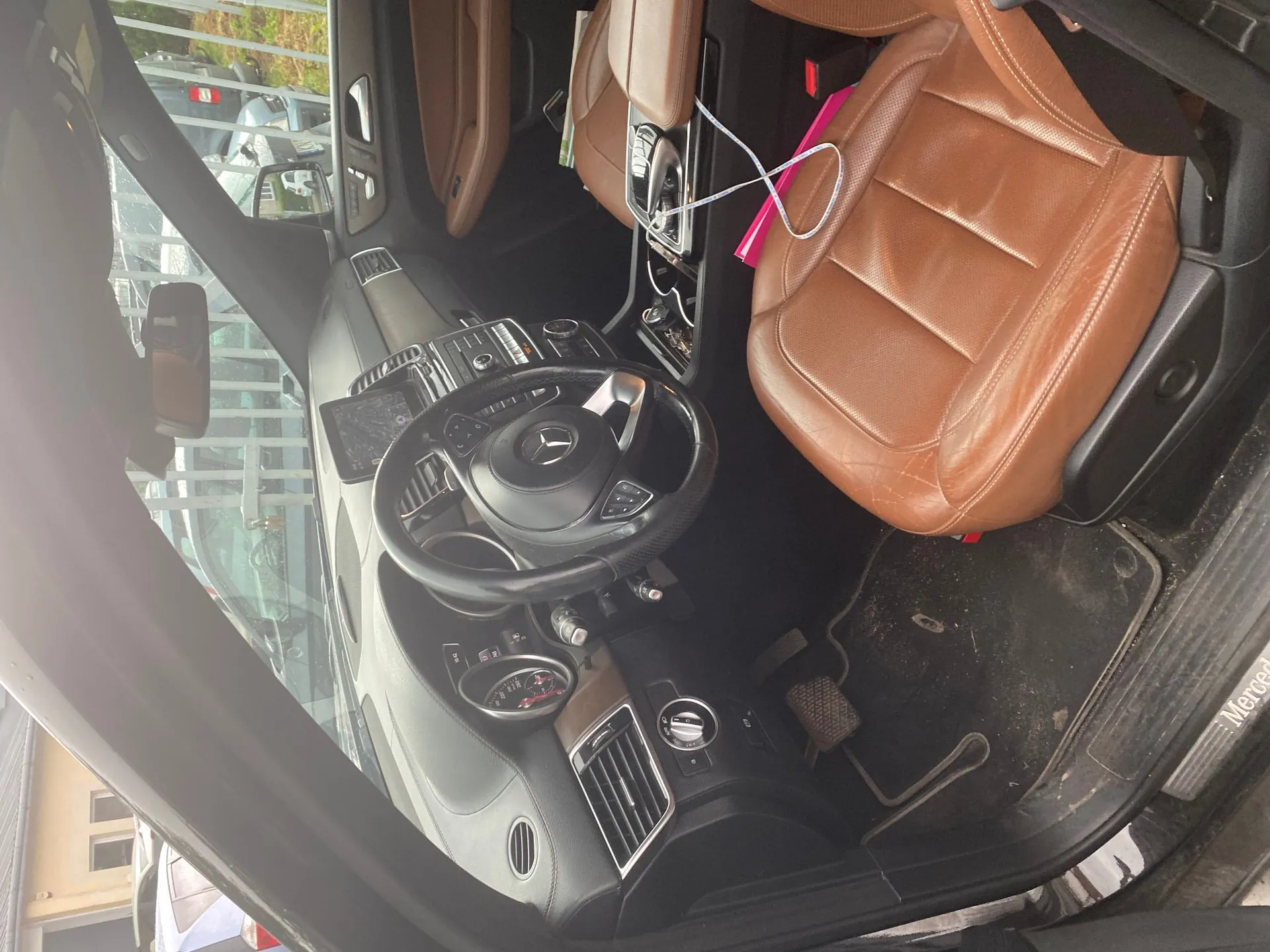 Wnętrze samochodu marki Mercedes-Benz z perspektywy kierowcy. Widoczna jest czarna kierownica z logo marki, deska rozdzielcza z przyciskami i ekranem oraz brązowe, skórzane fotele. Na siedzeniu pasażera leży różowy przedmiot i biały kabel. Na progu samochodu widoczny jest napis 'Mercedes'.