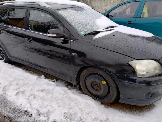 Czarny samochód osobowy z pokrytą śniegiem maską i przednią szybą, bez przedniego koła. Na tle widoczny turkusowy samochód. Śnieg na ziemi wokół pojazdów.