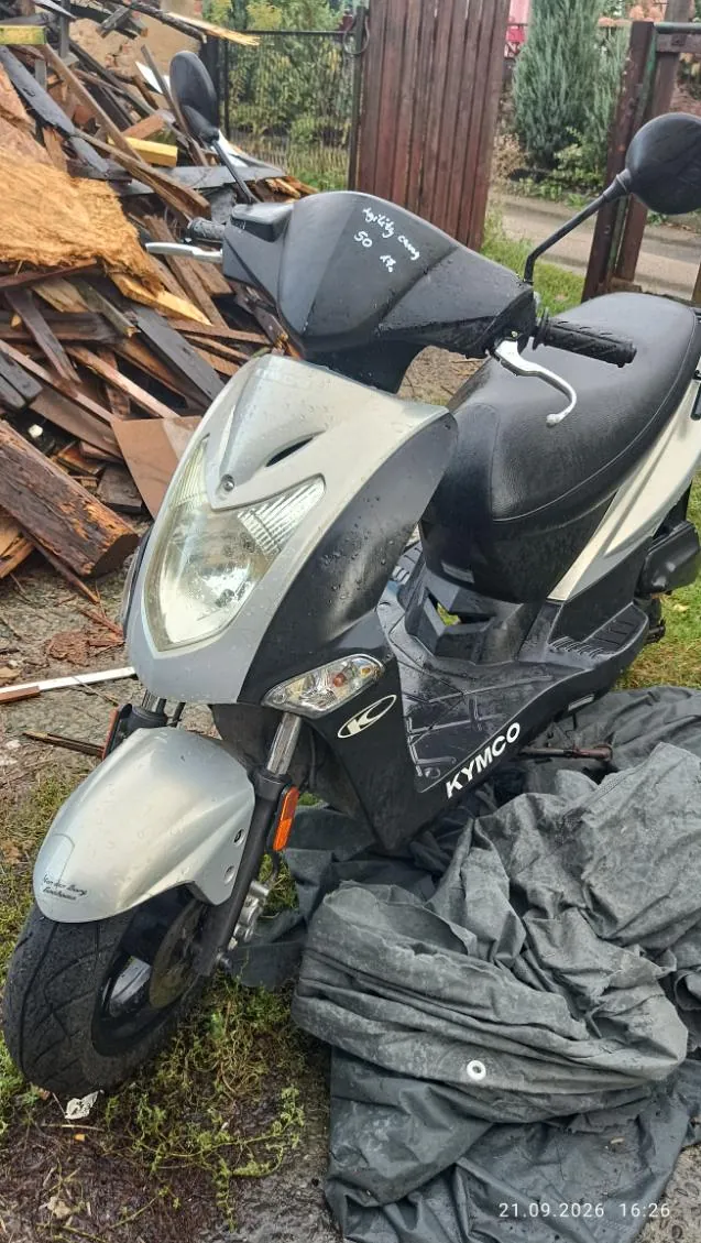 Srebrno-czarny skuter marki KYMCO zaparkowany na trawie obok sterty drewnianych desek i kawałków drewna. Skuter ma widoczny napis 'KYMCO' na boku oraz odręczny napis 'agility ceny 50 zł' na przedniej osłonie kierownicy. Pod skuterem leży szara plandeka. W prawym dolnym rogu zdjęcia znajduje się data i godzina: '21.09.2026 16:26'.