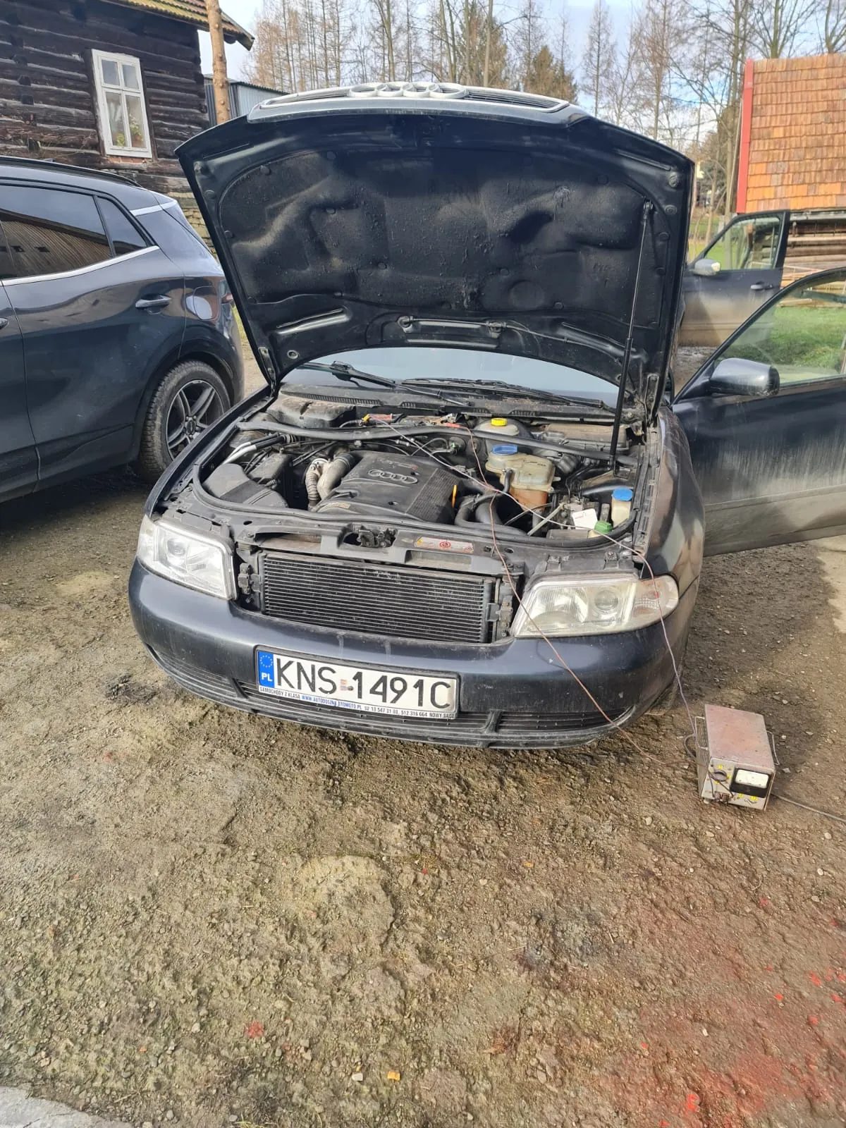 Czarny samochód marki Audi z otwartą maską, ukazującą silnik. Widoczna tablica rejestracyjna PL KNS 1491C. Po lewej stronie czarny SUV, po prawej samochód z otwartą drzwiami kierowcy. Tło: drewniane domy, drzewa bez liści, ziemia z drobnymi kamieniami. Na ziemi obok samochodu urządzenie elektryczne z przewodami.