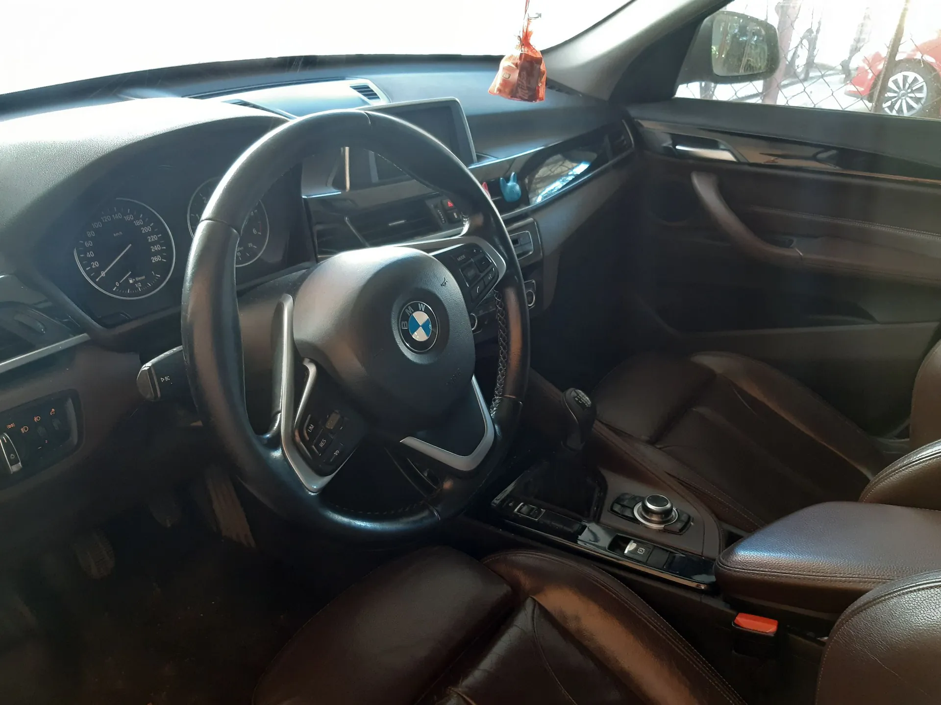 Wnętrze samochodu marki BMW z perspektywy kierowcy. Widoczna jest czarna skórzana kierownica z logo BMW w centrum, brązowe skórzane fotele oraz deska rozdzielcza z zegarami analogowymi. W tle widać fragmenty konsoli środkowej, lewarek zmiany biegów oraz lusterko wsteczne, w którym odbija się fragment otoczenia zewnętrznego.