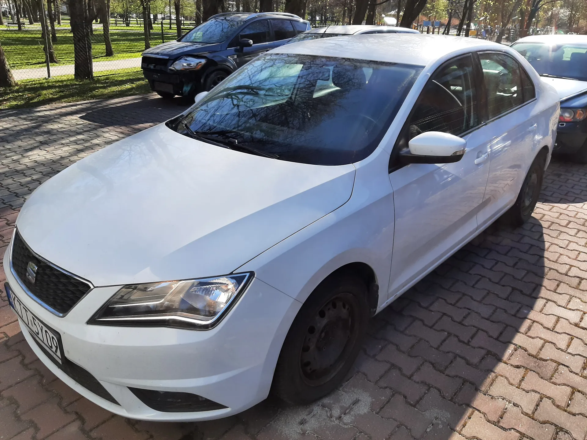 Biały samochód osobowy marki SEAT z tablicą rejestracyjną KT15Y09 zaparkowany na brukowanym placu. W tle czarny SUV z uszkodzonym przodem, niebieski samochód oraz drzewa, trawnik i ogrodzenie. Słoneczna pogoda, cienie rzucane na bruk.