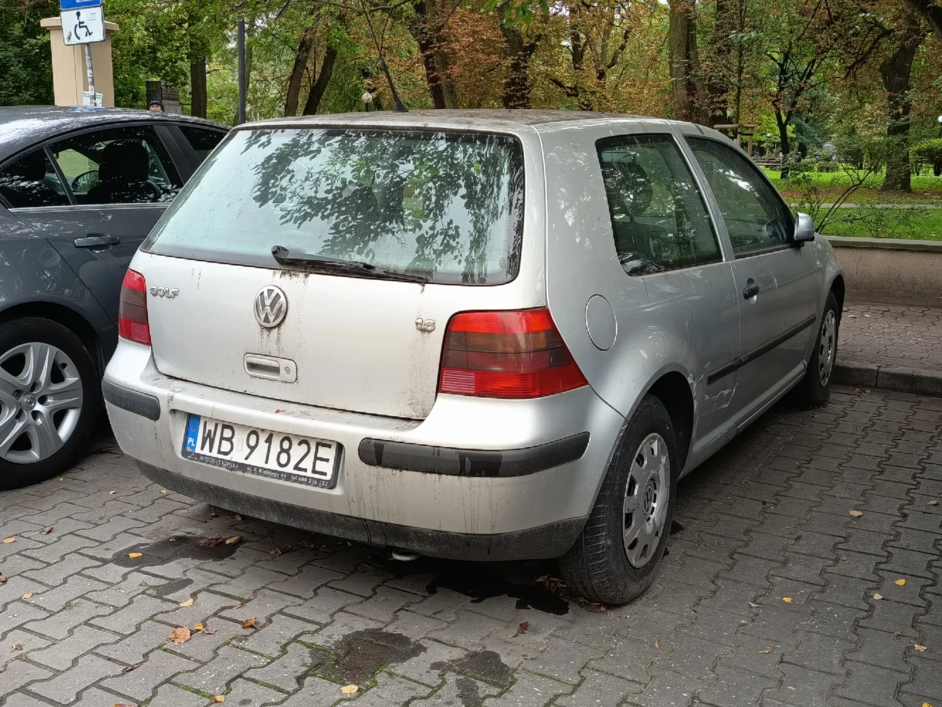 Srebrny samochód marki Volkswagen Golf zaparkowany na kostce brukowej. Widoczny jest tył i prawy bok pojazdu. Samochód jest zabrudzony, szczególnie w dolnej części klapy bagażnika. Tablica rejestracyjna ma numer WB 9182E. Po lewej stronie widoczny jest fragment innego, ciemnoszarego samochodu. W tle znajdują się zielone drzewa i krzewy.