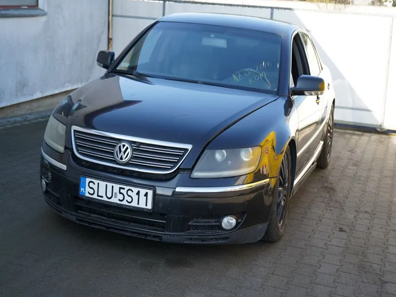 Czarny samochód marki Volkswagen z tablicą rejestracyjną SLU 5S11 (PL) zaparkowany na brukowanej powierzchni. Na przedniej szybie widoczny napis ręcznie zapisany: 12.04.2016 KOM. W tle biała ściana i metalowa brama. Samochód ma chromowaną kratę chłodnicy z logo VW oraz reflektory.