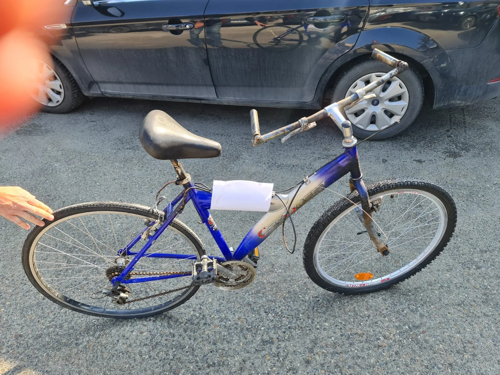 Niebieski rower z czarnym siedziskiem i rdzewnymi kierownicami, z białym arkuszem papieru przymocowanym do ramy. Rower stoi na asfalcie, w tle ciemny samochód osobowy. Widoczna ręka dotykająca tylnej opony roweru. Opony mają srebrne felgi, a na przedniej oponie widoczny pomarańczowy odblask.