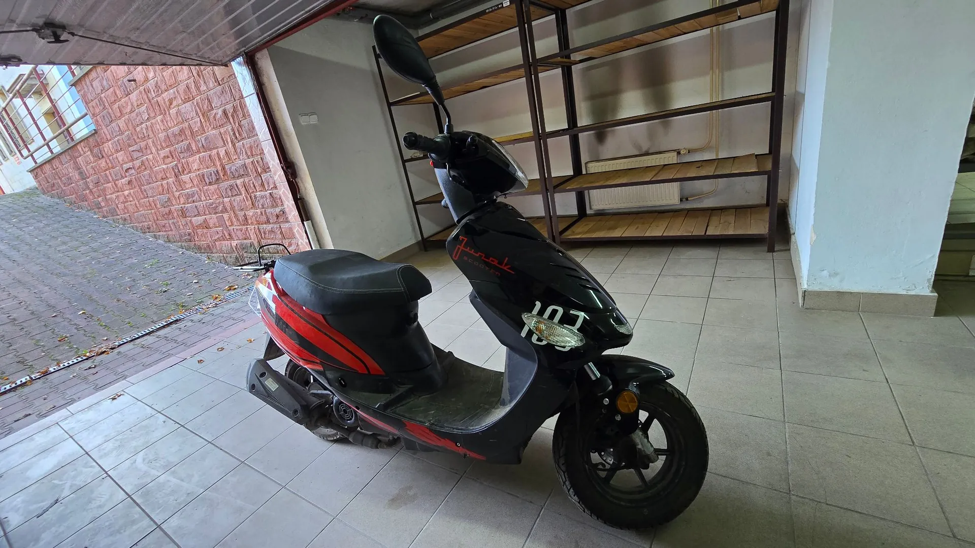 Czarny skuter z czerwonymi i pomarańczowymi pasami na boku, zaparkowany w garażu na jasnej, kafelkowej podłodze. Na przedniej owiewce widnieje napis 'Junak'. W tle znajdują się metalowe regały z drewnianymi półkami oraz ściana z czerwonej cegły po lewej stronie.