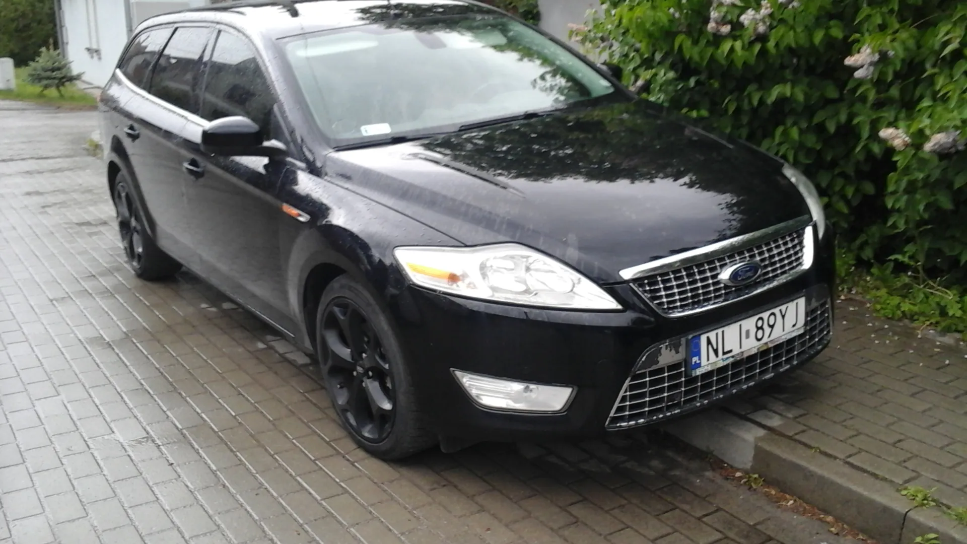 Czarny samochód osobowy typu kombi marki Ford zaparkowany na mokrej, brukowanej nawierzchni. Widoczna tablica rejestracyjna z polskim kodem PL i numerem NL 89YJ. Po prawej stronie zielone krzewy z kwiatami, po lewej budynek i trawnik. Samochód ma czarne felgi, reflektory i chromowaną atrapę chłodnicy z logo Ford.