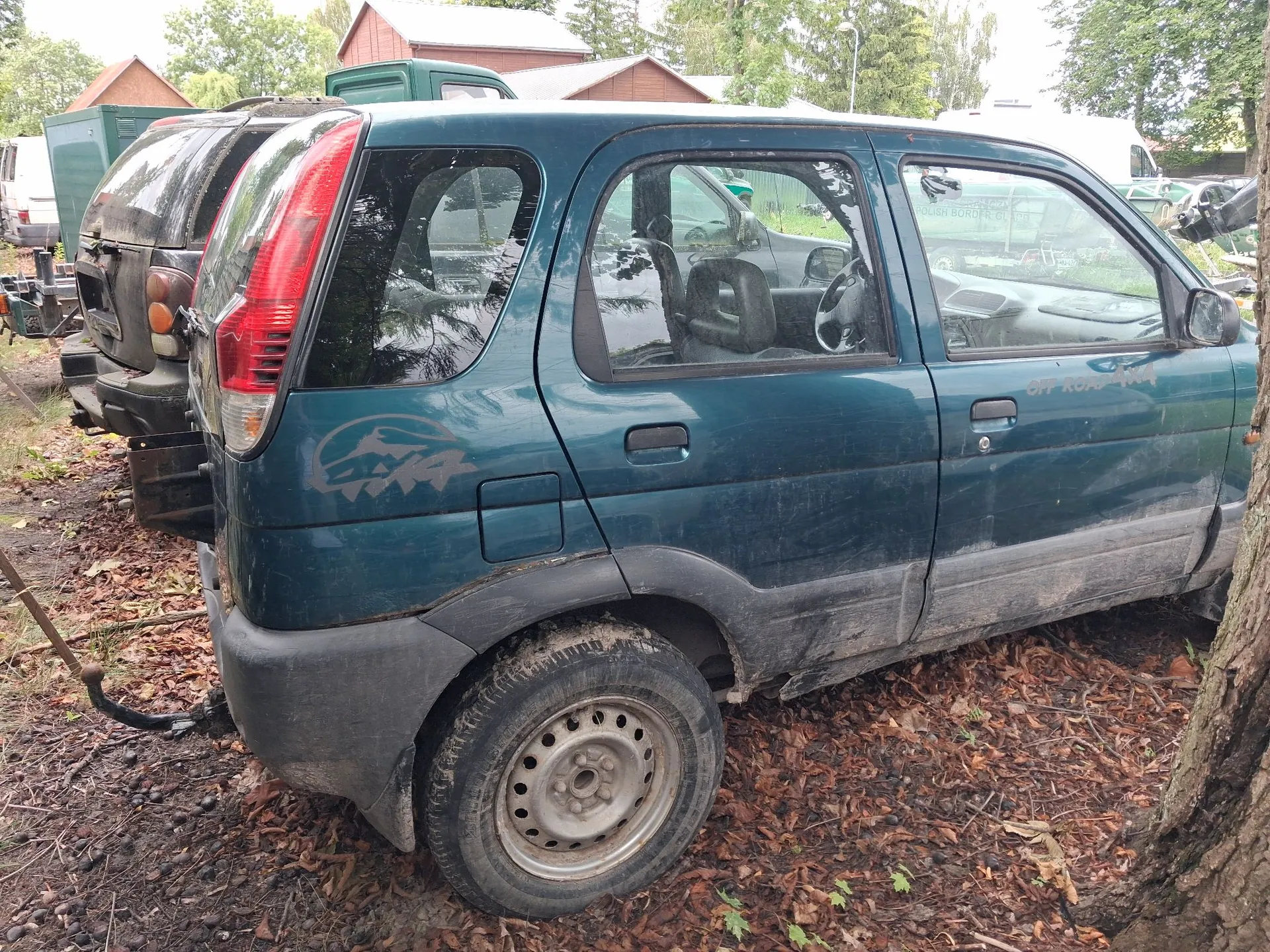 Widok z boku na ciemnozielony, zabrudzony samochód typu hatchback zaparkowany na ziemi pokrytej suchymi liśćmi. Na tylnym błotniku pojazdu znajduje się szara naklejka z sylwetką zwierzęcia i napisem. W tle widoczne są inne zaparkowane samochody oraz drewniane domki z czerwonymi dachami i drzewa.