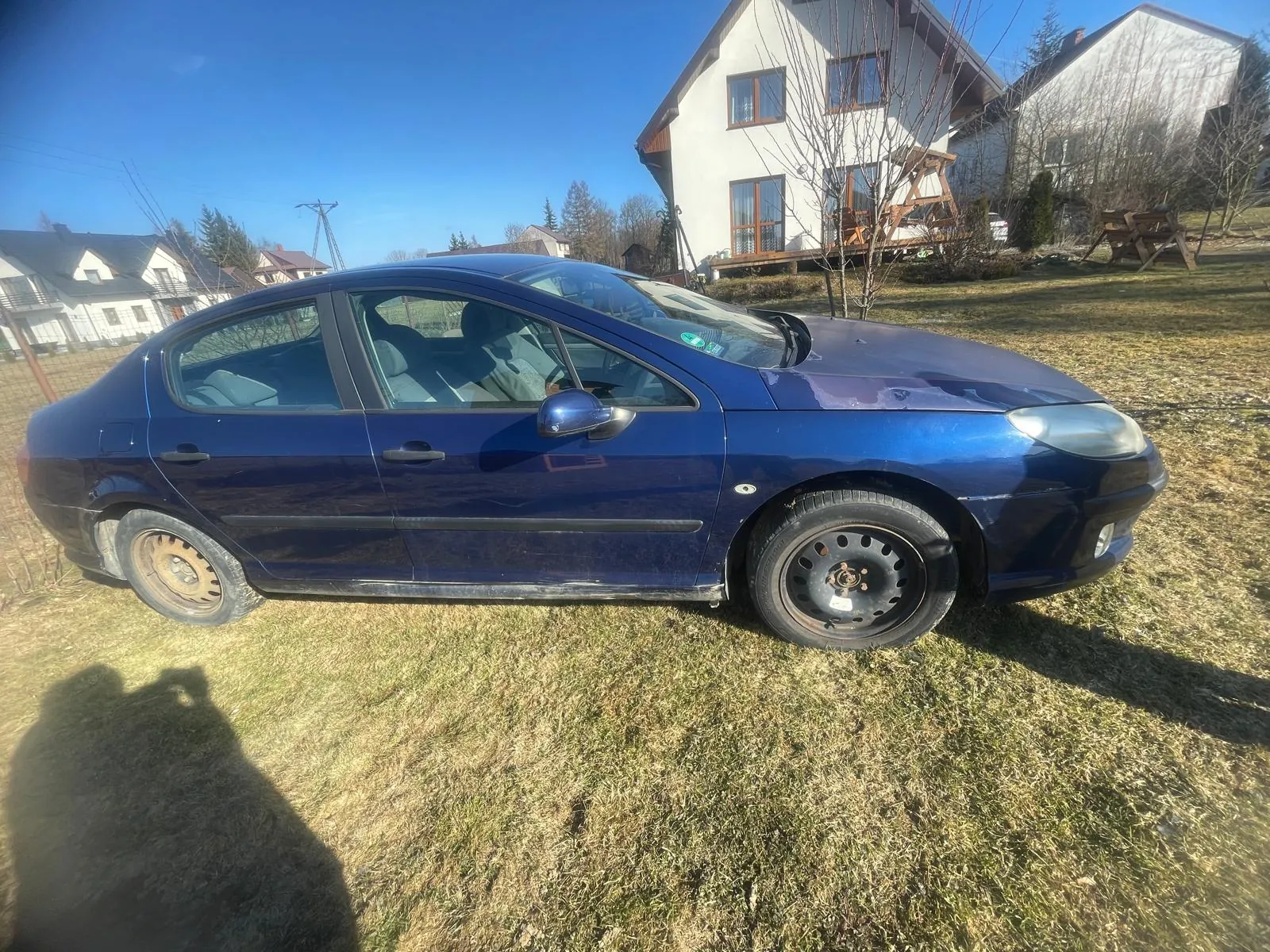 Niebieski samochód osobowy typu sedan zaparkowany na trawie. Pojazd ma czarne felgi, a na przednim szybie widoczny jest zielony znaczek. W tle znajdują się domy jednorodzinne, suche drzewa i jasne niebo. Na trawie widoczny jest cień osoby robiącej zdjęcie.