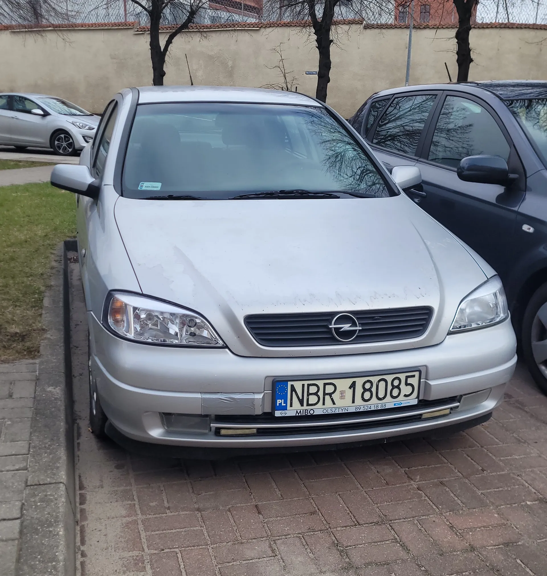 Srebrny samochód marki Opel zaparkowany na ceglanym chodniku. Widoczna tablica rejestracyjna PL NBR 18085 z napisem MIBO OLSZTYN oraz numerem telefonu 89 524 18 88. Po prawej stronie ciemnoszary samochód, w tle srebrny samochód, trawa, chodnik, beżowa ściana i drzewa bez liści. Na przedniej szybie naklejka.