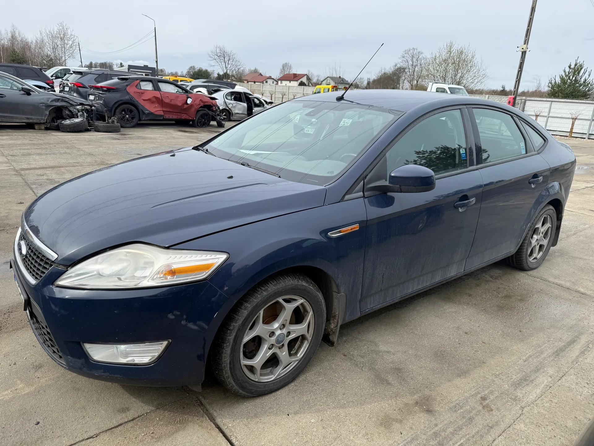 Ciemnoniebieski samochód osobowy typu sedan zaparkowany na betonowym placu. W tle widoczne są uszkodzone pojazdy, w tym czerwony i czarny samochód z deformacjami nadwozia, rozrzucone opony oraz ogrodzenie. Za ogrodzeniem znajdują się domy z czerwonymi dachami, drzewa i słup telekomunikacyjny. Niebo jest zachmurzone.