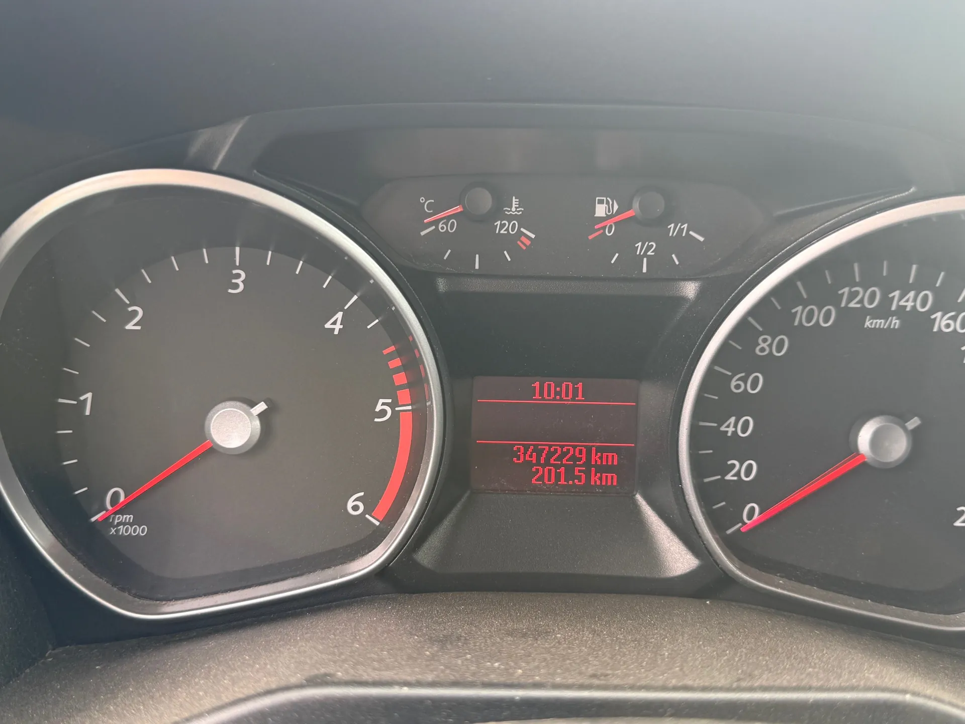 Zbliżenie na deski rozdzielcze samochodu. Po lewej stronie wskaźnik obrotomieru z zakresem 0-6 (oznaczony jako rpm x1000), po prawej wskaźnik prędkości z zakresem 0-160+ km/h. W centralnej części wyświetlacz cyfrowy z czerwonymi cyframi: godzina 10:01, przebieg 347229 km oraz 201.5 km. Powyżej wyświetlacz znajdują się wskaźniki temperatury chłodniczki (60-120°C) i poziomu paliwa (0-1/1). Wszystkie wskaźniki mają czarne tło z srebrnymi obramowaniami i czerwonymi wskazówkami.