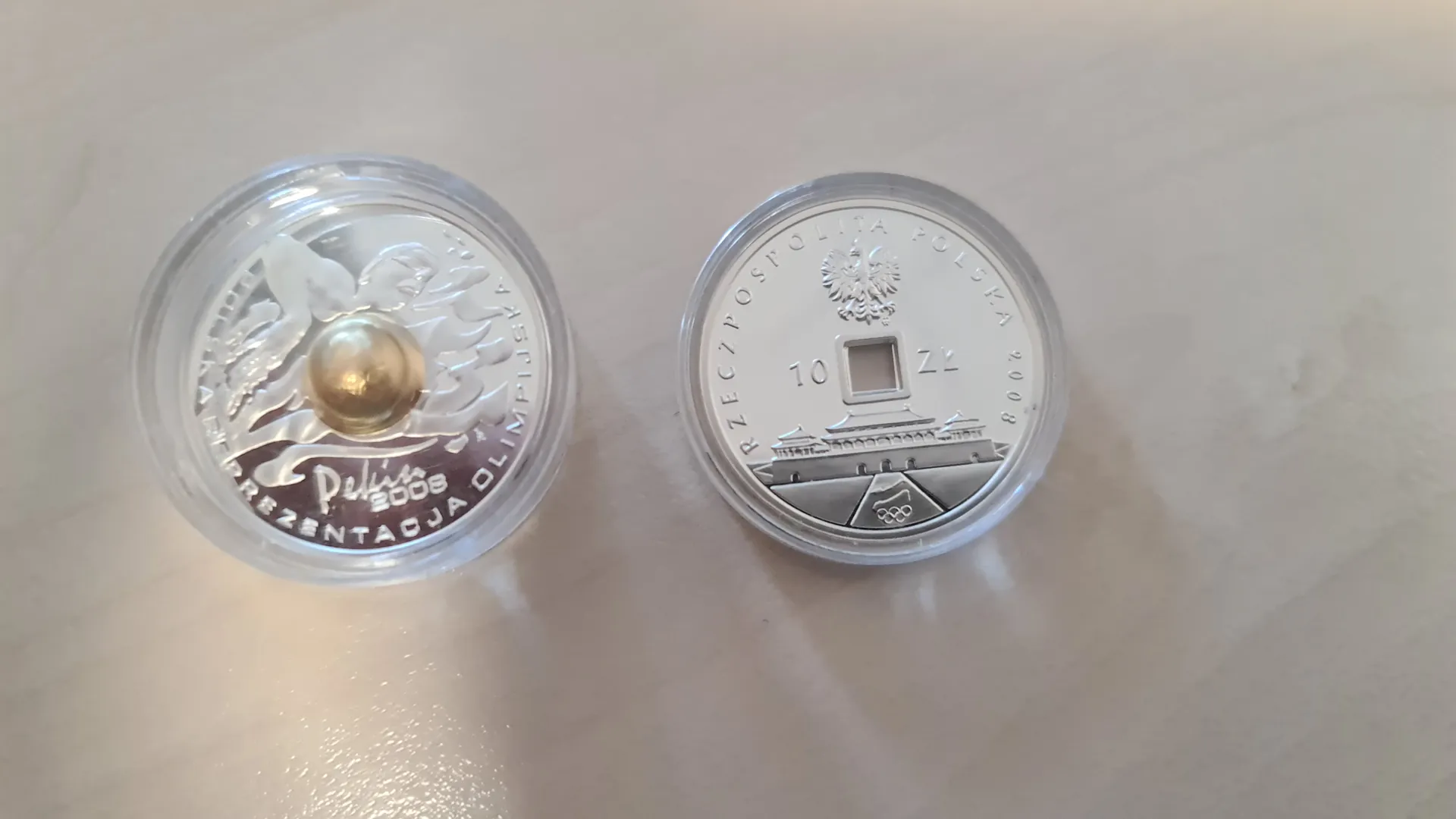 Dwa srebrne monety w przezroczystych etui leżące na jasnej powierzchni. Lewa moneta przedstawia anioła z rozpostartymi skrzydłami i złotym okrągłym elementem w centrum, z napisem 'Pekin 2008' oraz 'REPREZENTACJA OLIMPIJSKA'. Prawa moneta ma orła w koronie, napis 'RZECZPOSPOLITA POLSKA', wartość '10 ZŁ', rok '2008', przedstawienie budynku i pierścienie olimpijskie.
