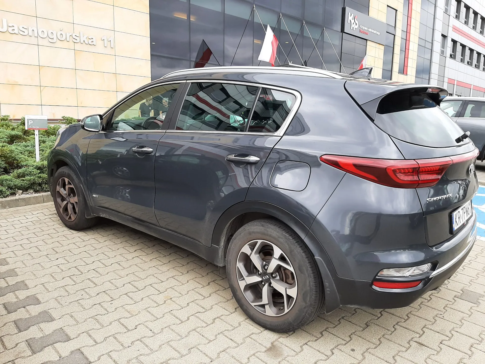 Szary samochód marki Kia Sportage zaparkowany na kostce brukowej przed nowoczesnym budynkiem. Na ścianie budynku widnieje napis 'Jasnogórska 11'. W tle widoczne są szklane witryny oraz dwie flagi, w tym biało-czerwona flaga Polski.