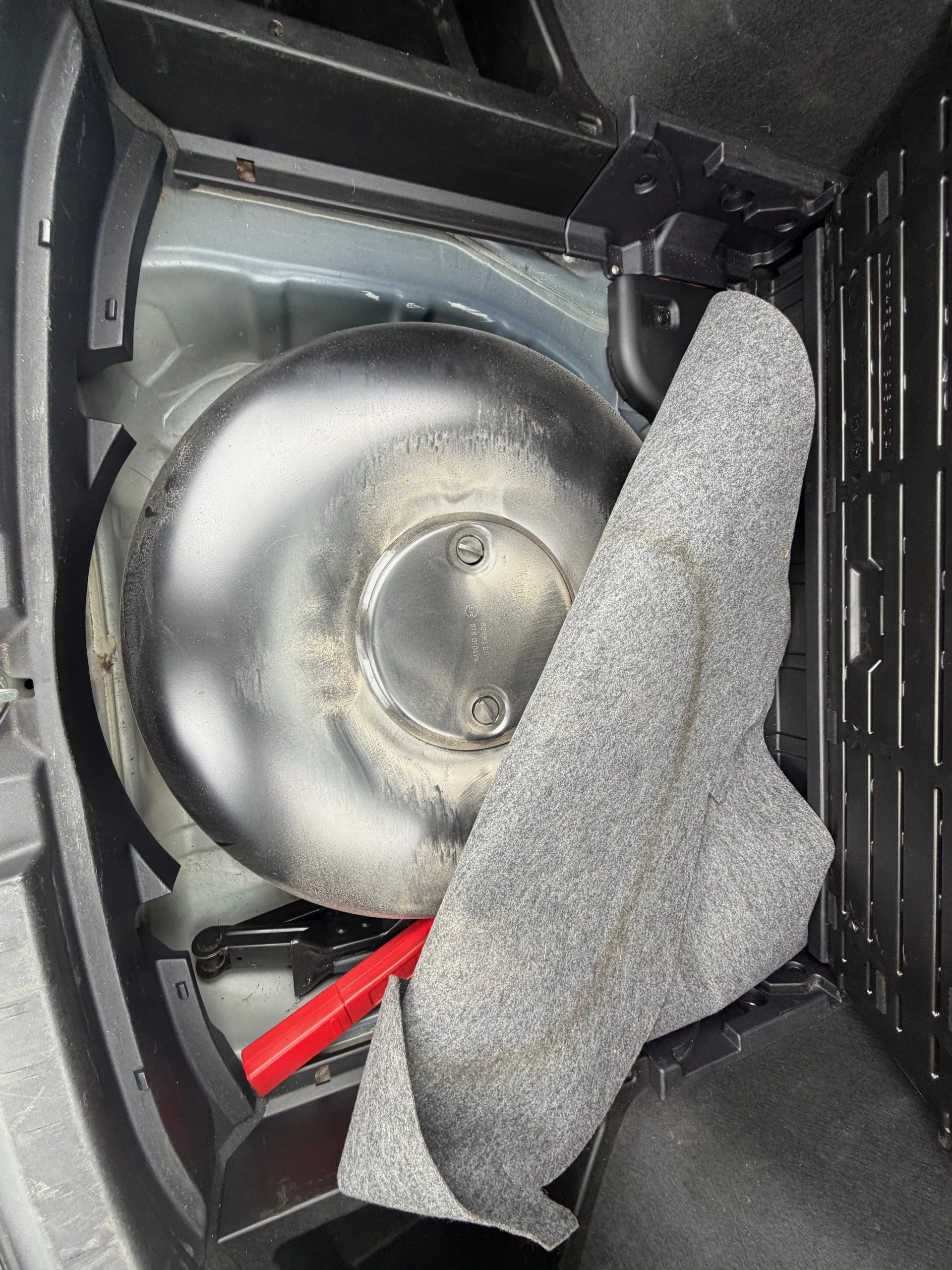 Wnętrze bagażnika samochodu z umieszczonym w środku kołem zapasowym o metalowej powierzchni. Na kole widoczne są dwa śruby i nieczytelny napis. Przy kolu leży szara tkanina oraz czerwony przedmiot przypominający klucz lub podnośnik. Wokół znajdują się czarne plastikowe elementy konstrukcyjne bagażnika.