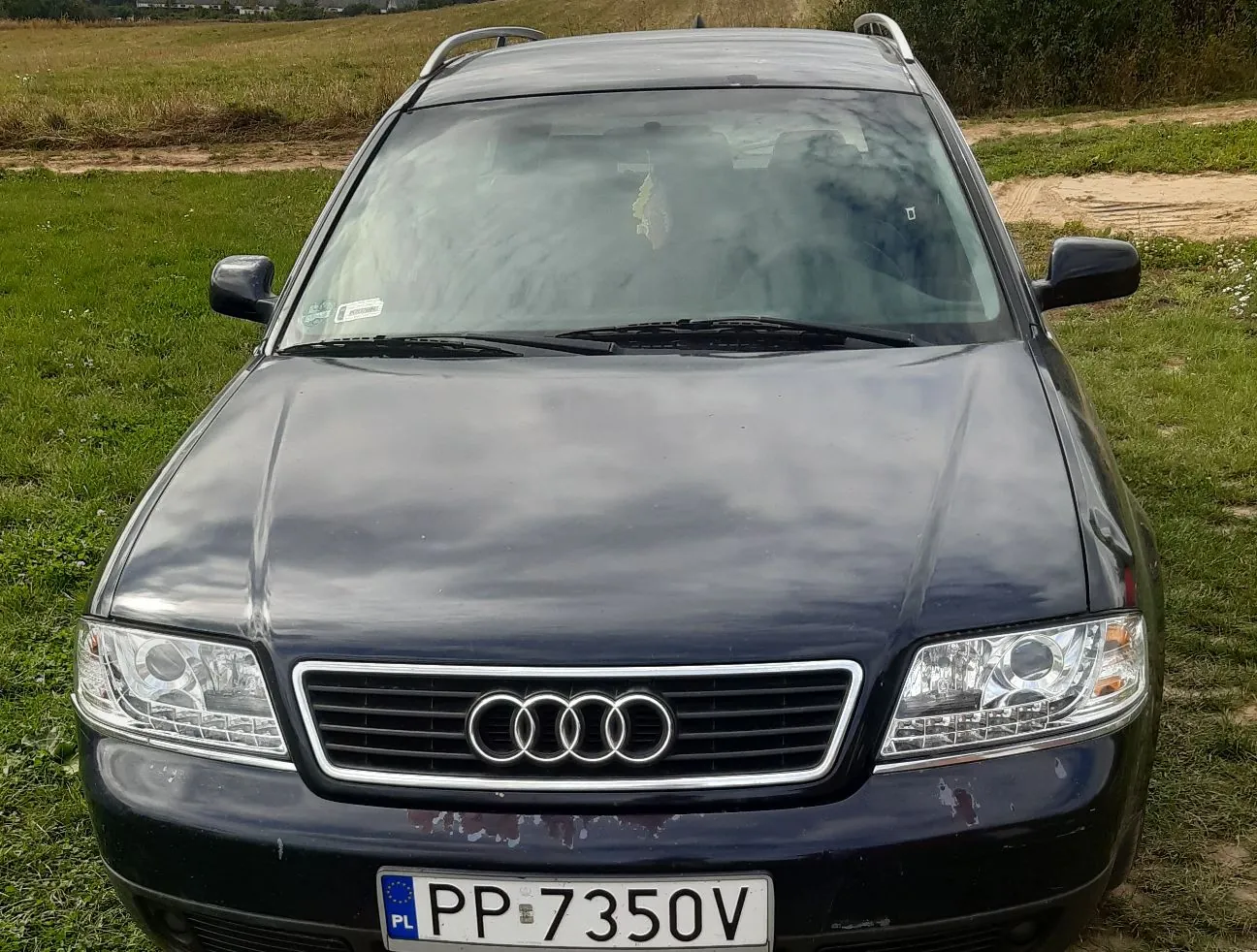 Czarny samochód marki Audi zaparkowany na trawiastym terenie. Widoczny jest przód pojazdu z charakterystycznym grillem i logo czterech pierścieni. Samochód posiada relingi dachowe. Tablica rejestracyjna ma numer PP 7350V i oznaczenie PL.