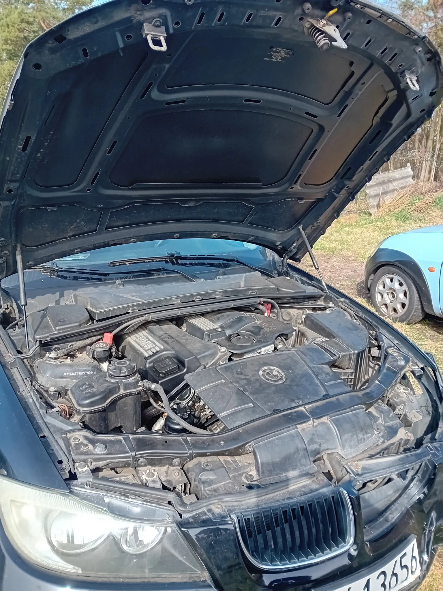 Czarny samochód marki BMW z otwartą maską, ukazujący silnik z widocznym logo BMW. W tle niebieski samochód zaparkowany na trawie oraz drzewa. Tablica rejestracyjna z częściowo widocznymi cyframi 13656.