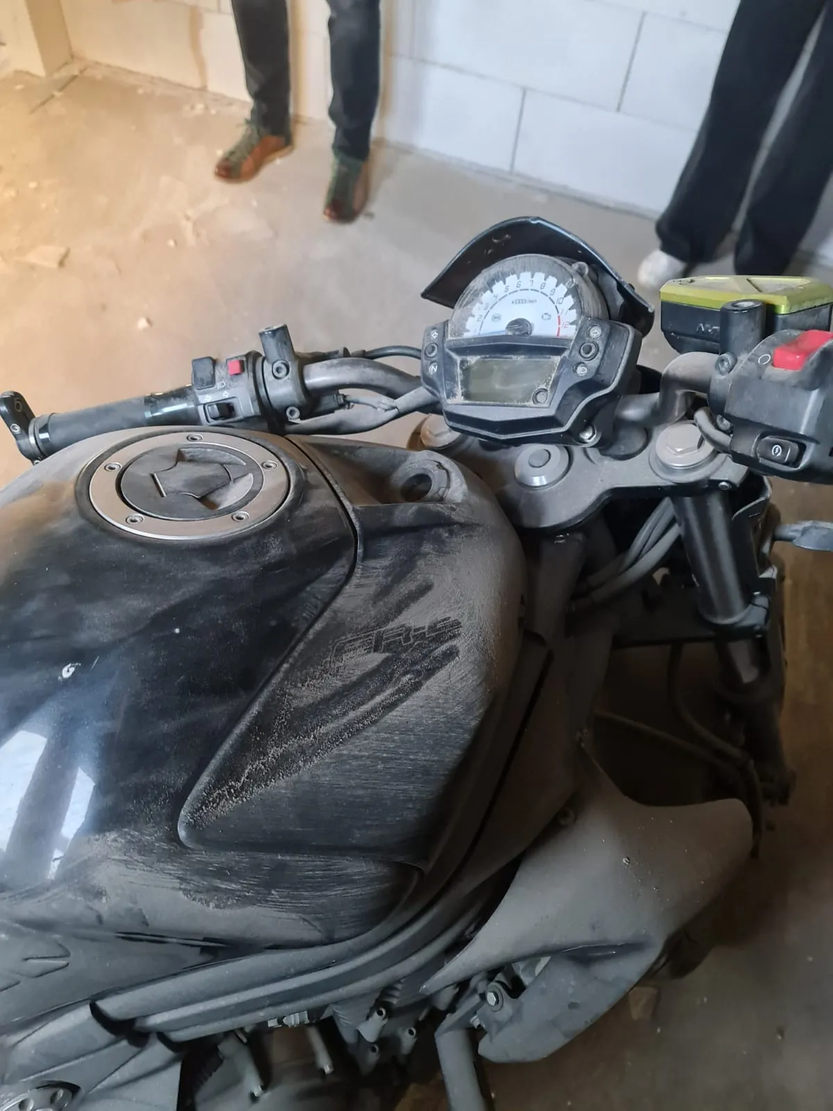 Czarny motocykl z widocznym zbiornikiem paliwa, kierownicą i licznikiem prędkości. Zbiornik pokryty jest kurzem, na nim widoczny jest metalowy element z otworem w kształcie gwiazdy. Licznik prędkości ma białą tarczę z czarnymi cyframi i czerwoną igłą. W tle widoczne są nogi dwóch osób w czarnych spodniach i butach, betonowa podłoga oraz biała ściana z kafelkami.