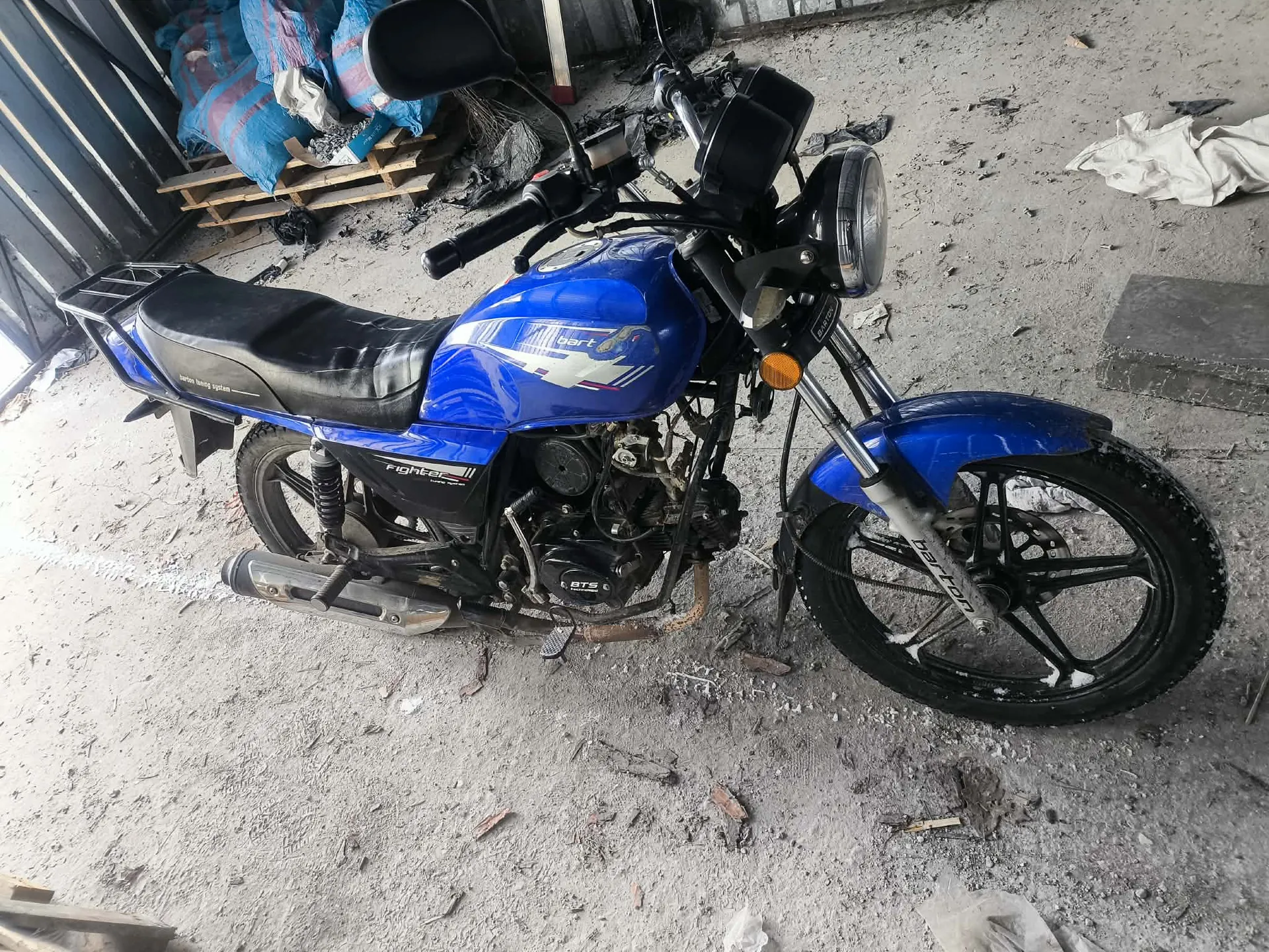 Niebieski motocykl z czarnym siedzeniem i widocznymi elementami silnika. Na przednim kole widoczny napis 'Bajaj'. Motocykl znajduje się na betonowej powierzchni z rozrzuconymi śmieciem i fragmentami materiałów. W tle widoczne są niebiesko-fioletowe worki na drewnianych paletach oraz metalowa konstrukcja.