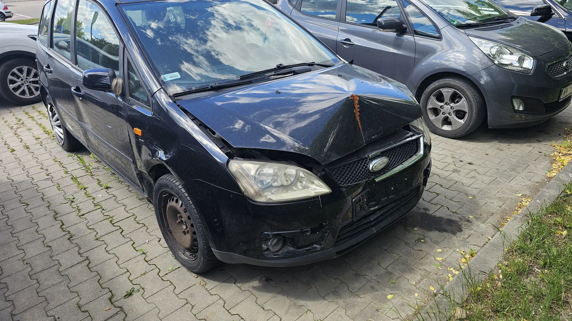 Czarny samochód osobowy z widocznymi uszkodzeniami przedniej części: zgnieciona maska z rdzawą plamą po prawej stronie, uszkodzony zderzak i reflektor. Pojazd zaparkowany na brukowanym placu parkingowym z trawą i chwastami między płytami. W tle widoczne są inne samochody: szary i srebrny. Na przedniej szybie naklejka. Niebo odbijające się w szybie jest częściowo zachmurzone.