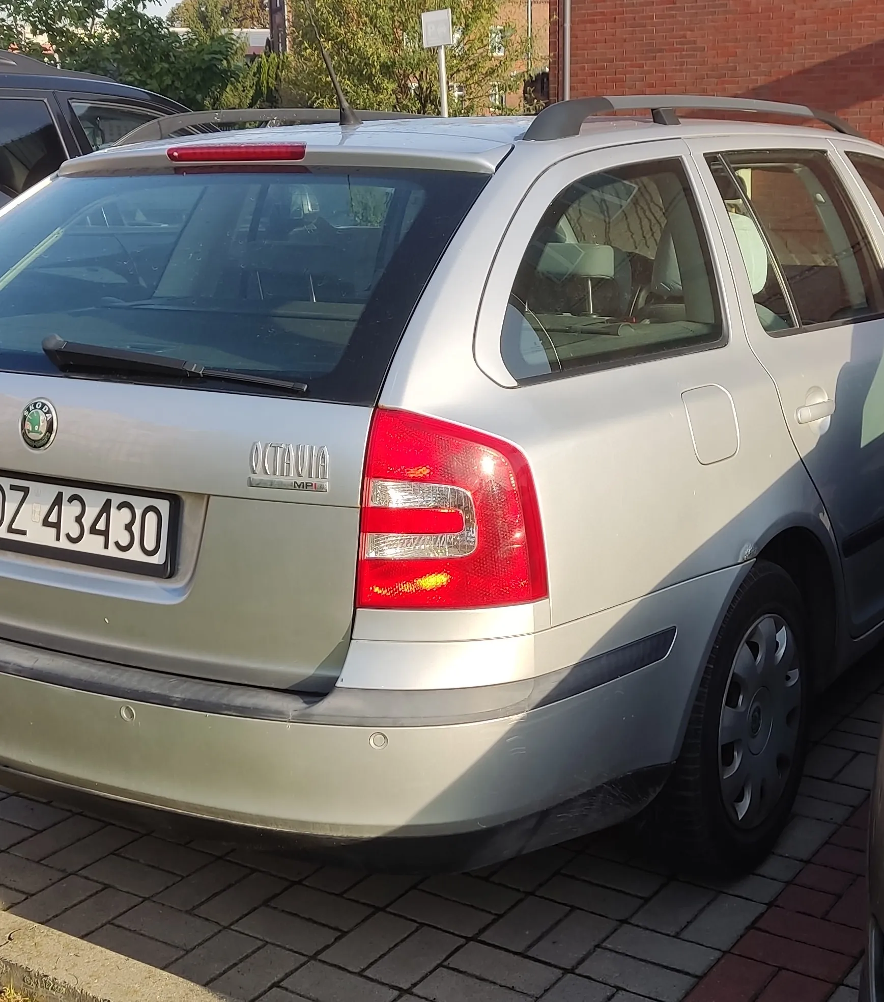 Srebrny samochód marki Skoda Octavia Combi zaparkowany na kostce brukowej. Widoczny jest tył pojazdu z lewej strony, tablica rejestracyjna o numerze DZ 43430 oraz logo marki i napis OCTAVIA. Samochód posiada relingi dachowe.