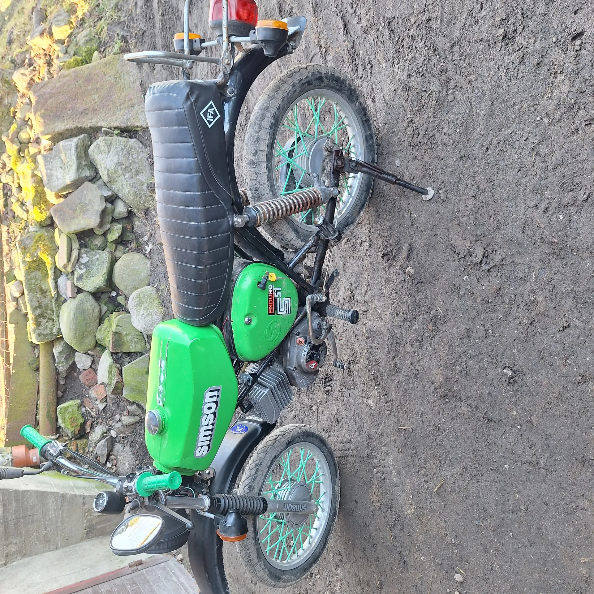 Zielony motocykl marki Simson z czarnym siedzeniem i logo IFA, położony na boku na brudnej ziemi. Koła z zielonymi promieniami, zielony zbiornik paliwa z napisem 'Simson' oraz czarne elementy konstrukcyjne. W tle kamienie i mchy.