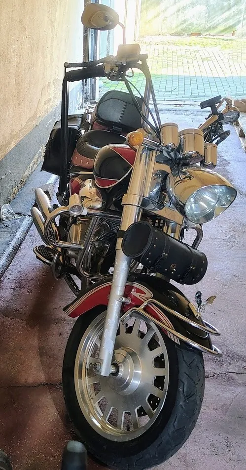 Zdjęcie motocykla typu cruiser zaparkowanego w garażu lub podwórzu. Motocykl ma chromowane elementy, w tym przedni widelec, reflektor i rury wydechowe. Posiada czarną skórzaną kanapę z czerwonymi akcentami oraz czarny, cylindryczny kufer zamontowany z przodu. Przednie koło ma srebrną, wieloramienną felgę.