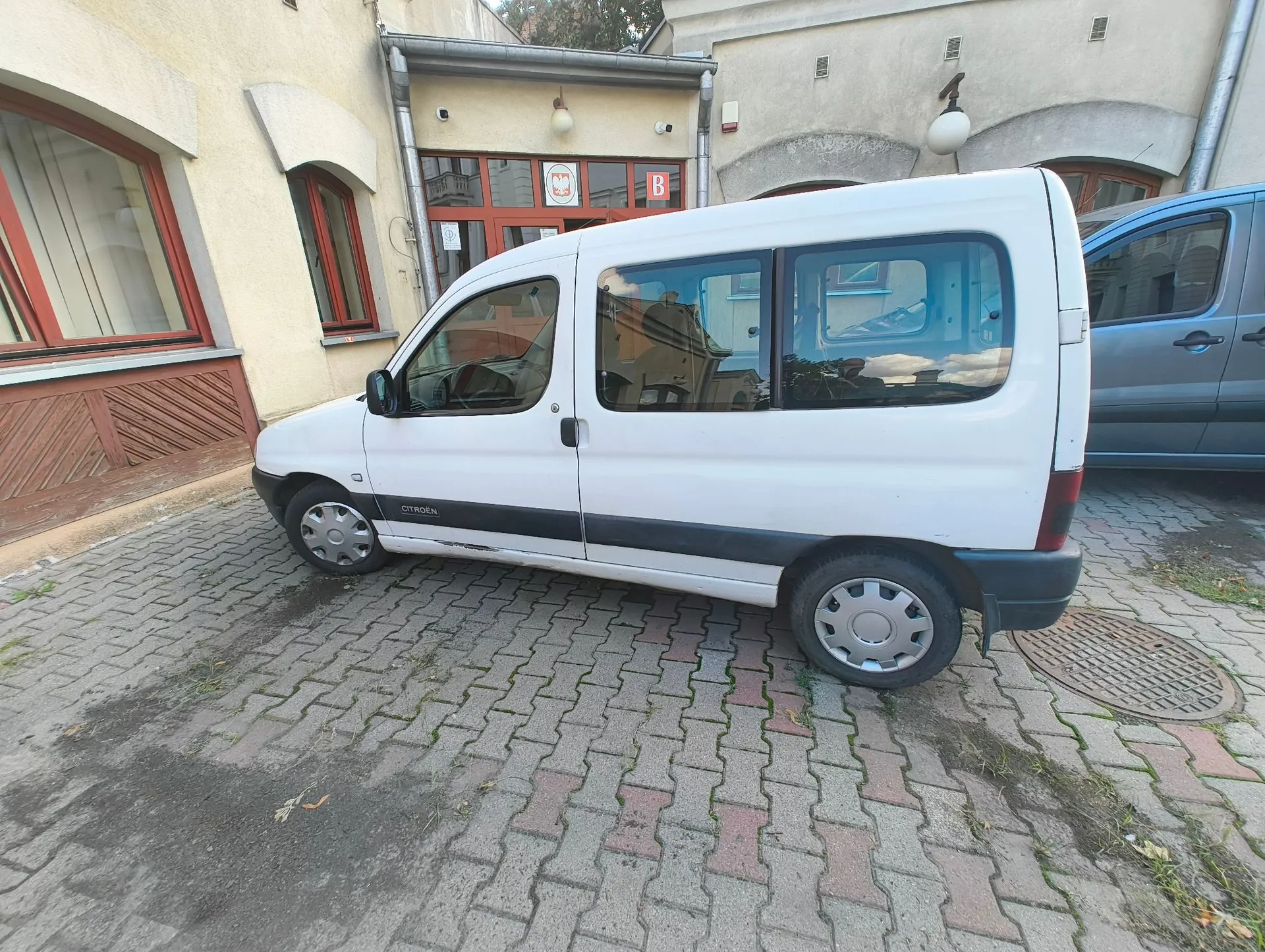 Biały samochód dostawczy marki Citroën zaparkowany na kostce brukowej przed budynkiem. Pojazd jest ustawiony bokiem do widza. W tle widoczny jest jasny budynek z oknami i wejściem, nad którym znajduje się czerwona tabliczka z literą 'B'. Obok białego vana stoi fragment innego, niebieskiego samochodu.