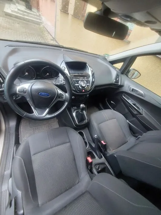 Wnętrze samochodu marki Ford widziane z perspektywy kierowcy. Widoczna jest czarna kierownica z logo Forda, deska rozdzielcza z ekranem multimedialnym, dwa przednie fotele z ciemnoszarej tkaniny oraz lewarek zmiany biegów. Obraz jest obrócony o 90 stopni w lewo.