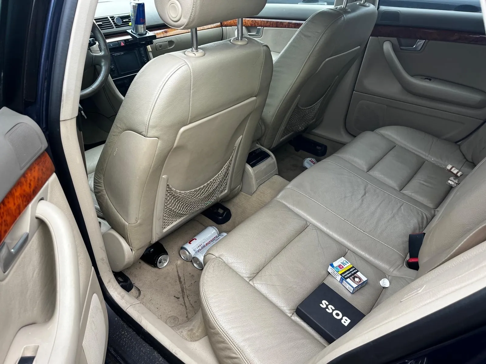 Wnętrze samochodu z beżowymi skórzany siedzeniami. Widoczne są przednie i tylne siedzenia, kierownica, deska rozdzielcza z ekranem oraz drewnianą obudową. Na tylnym siedzeniu leżą czarna pudełko z napisem "BOSS", paczka papierosów i mały przedmiot. Na podłodze znajdują się puste puszki, w tym jedna z czerwonym logo. W siatce na oparciu przedniego siedzenia widoczna jest pusta puszka. Drzwi samochodu są otwarte, pokazując uchwyt i głośnik.