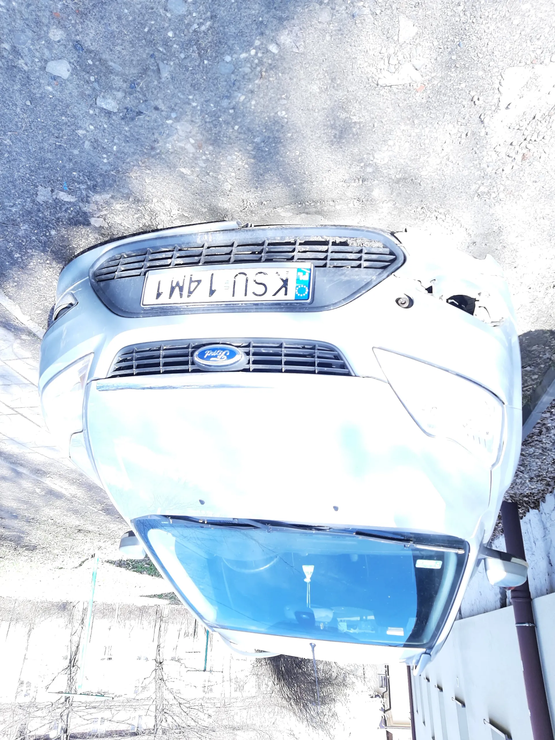 Samochód marki Ford w kolorze srebrnym z widoczną tablicą rejestracyjną KSU 14M1. Przednia prawa strona pojazdu uszkodzona. Samochód zaparkowany na drodze z żwirem. W tle budynki, suche drzewa i fragment drogi.