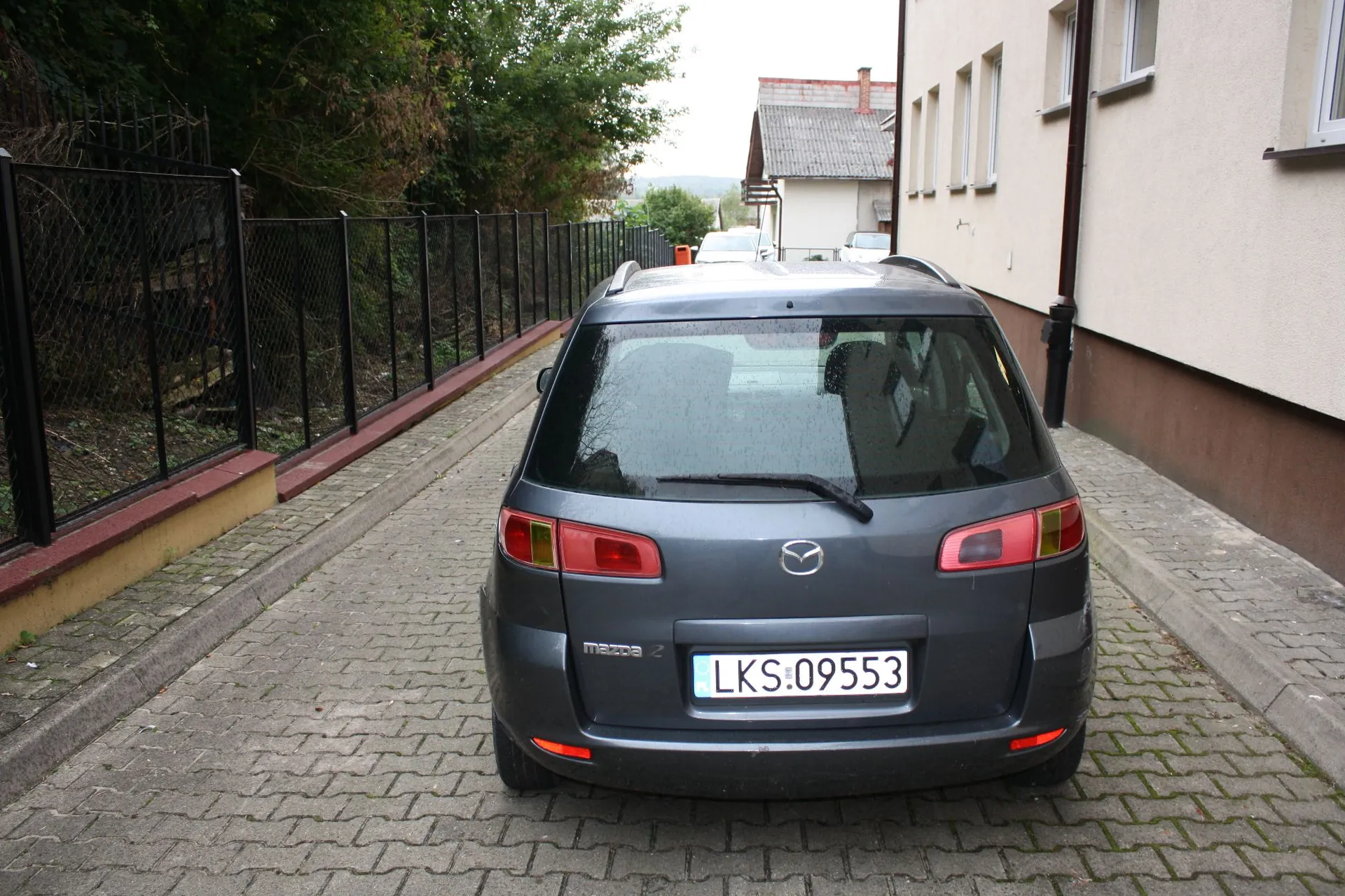 Srebrnoszary samochód marki Mazda zaparkowany tyłem na kostce brukowej. Pojazd posiada tablicę rejestracyjną o numerze LKS:09553. Po lewej stronie znajduje się czarny metalowy płot, a po prawej jasna ściana budynku. W tle widoczny jest inny dom oraz zieleń drzew.