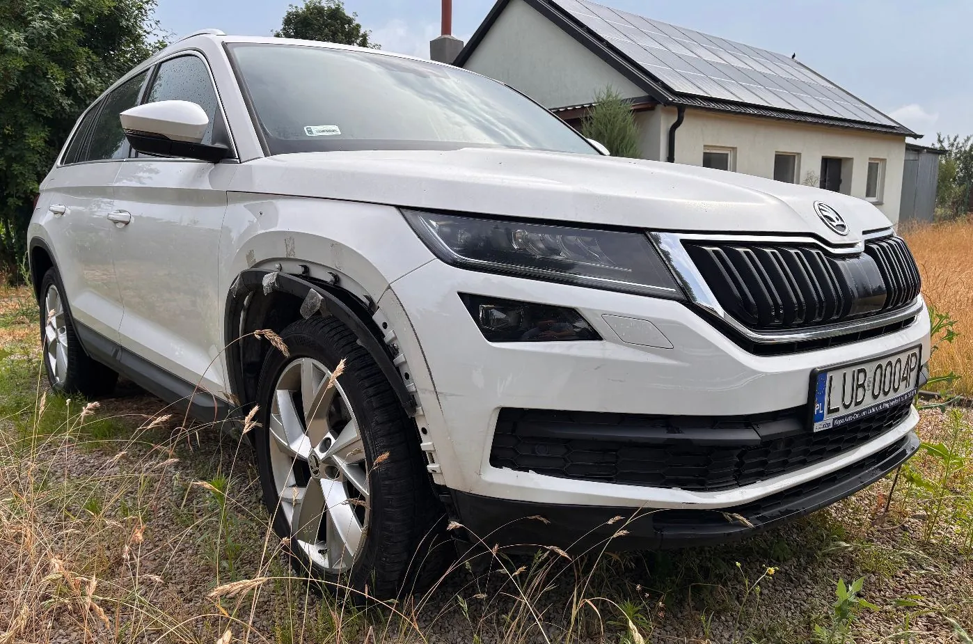 Biały samochód marki Škoda z widocznymi znacznymi uszkodzeniami przodu i prawego przedniego koła, które jest całkowicie zmiażdżone. Pojazd stoi na trawiastym terenie na tle białego domu z panelami fotowoltaicznymi na dachu.