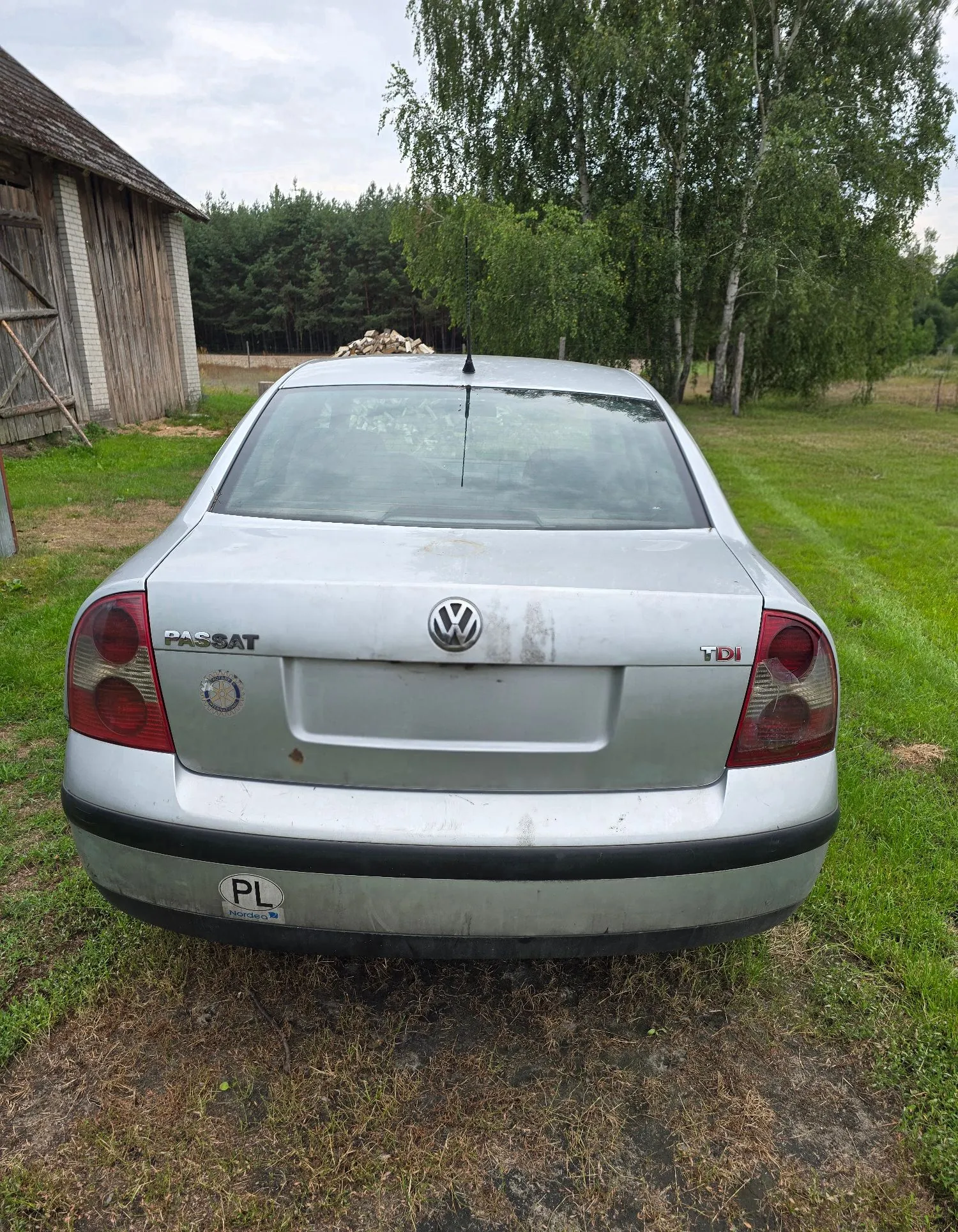 Srebrny samochód marki Volkswagen Passat TDI widziany od tyłu, zaparkowany na trawie przed drewnianą szopą i drzewami. Na klapie bagażnika widoczne są napisy 'PASSAT' oraz 'TDI' oraz logo Volkswagena. Na zderzaku znajduje się naklejka z oznaczeniem kraju 'PL'.