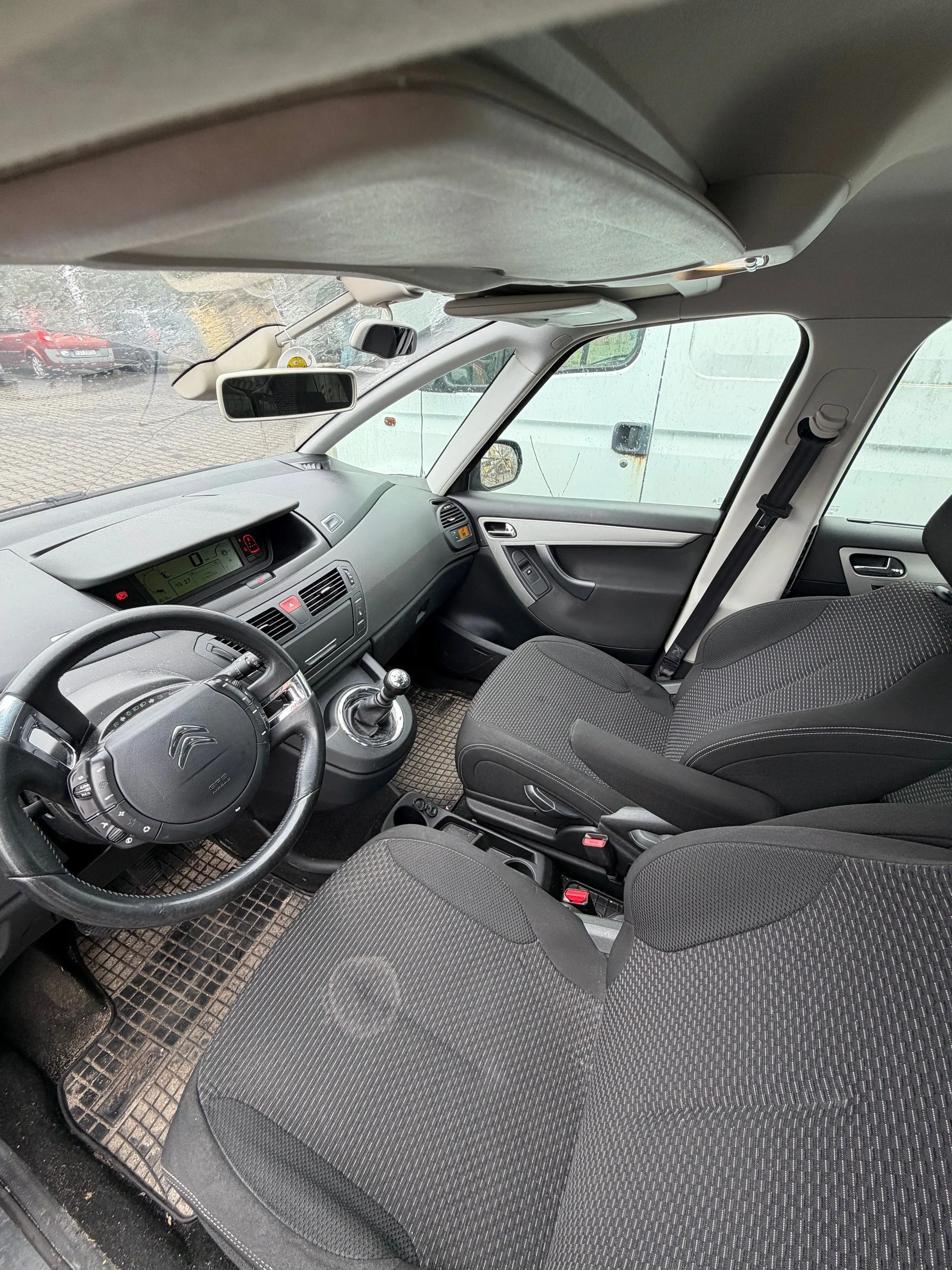 Wnętrze samochodu Citroën z widocznym kierownicą z logo marki, deska rozdzielcza z cyfrowym wyświetlaczem, dźwignią zmiany biegów, czarnymi siedzeniami w tkaninie z geometrycznym wzorem oraz dywanikami podłogowymi w kratkę. Przez przednią szybę widoczny czerwony samochód i budynek na zewnątrz. Lusterko wsteczne oraz klamki drzwi są w pozycji pionowej.