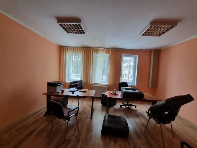 Pomieszczenie biurowe z pomarańczowymi ścianami i podłogą z drewnianych desek. Wnętrze zawiera dwa biurka z czarnymi i brązowymi krzesłami biurowymi. Na lewym biurku znajduje się drukarka oraz dokumenty, na prawym czerwony przedmiot. W tle trzy okna z żółtymi zasłonami, a na suficie dwa kwadratowe oprawy oświetleniowe. W pomieszczeniu widoczne są również czarne fotele i puf.