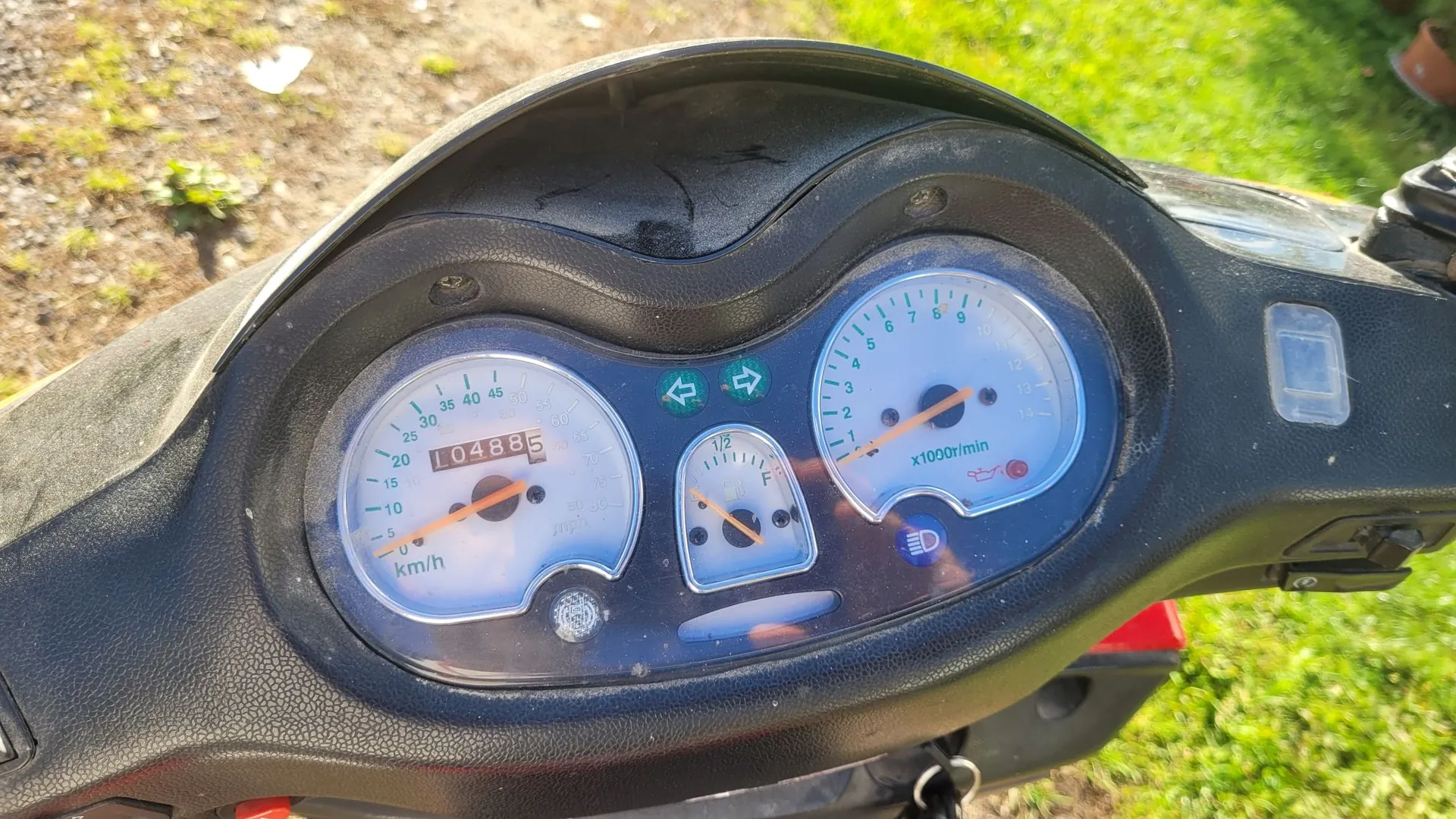 Zbliżenie na deskę rozdzielczą skutera. Widoczny jest prędkościomierz z zaznaczoną prędkością 0 km/h i licznikiem przebiegu wskazującym 10488,5 km, wskaźnik poziomu paliwa oraz obrotomierz z napisem x1000r/min. Pomiędzy wskaźnikami znajdują się kontrolki kierunkowskazów i świateł.