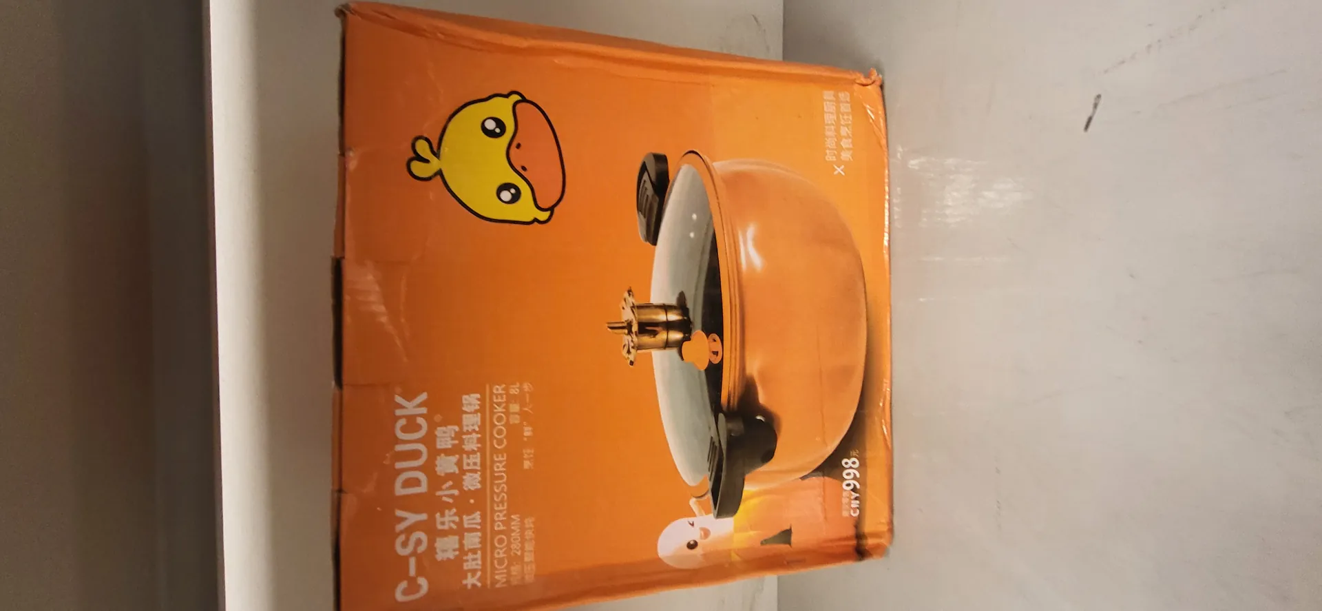 Pomarańczowe pudełko produktu z napisem 'C-SY DUCK' oraz chińskimi znakami '禧乐小黄鸭' i '大肚南瓜·微压料理锅'. Poniżej angielski opis 'MICRO PRESSURE COOKER', wymiary '280MM' i pojemność '8L'. Po prawej stronie ilustracja żółtego kaczora z pomarańczowym dziobem. W centralnej części obrazek pomarańczowego garnka ciśnieniowego z przezroczystą pokrywką i czarnymi uchwytami. W lewym dolnym rogu cena 'CNY 998', a po prawej dodatkowe chińskie napisy '时尚料理厨具 美食烹饪首选'. W lewym dolnym rogu mała postać w pomarańczowym stroju.