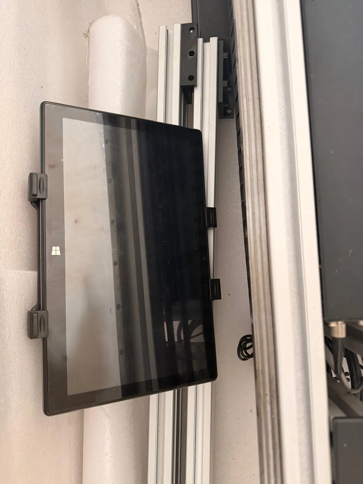 Czarny tablet lub panel dotykowy z włączonym ekranem, zamocowany do aluminiowego profilu konstrukcyjnego za pomocą dwóch czarnych uchwytów po lewej stronie. Na ekranie widoczne jest logo systemu Windows w lewym dolnym rogu. Obraz jest zorientowany pionowo, ale elementy konstrukcyjne sugerują, że urządzenie jest zamontowane bokiem.