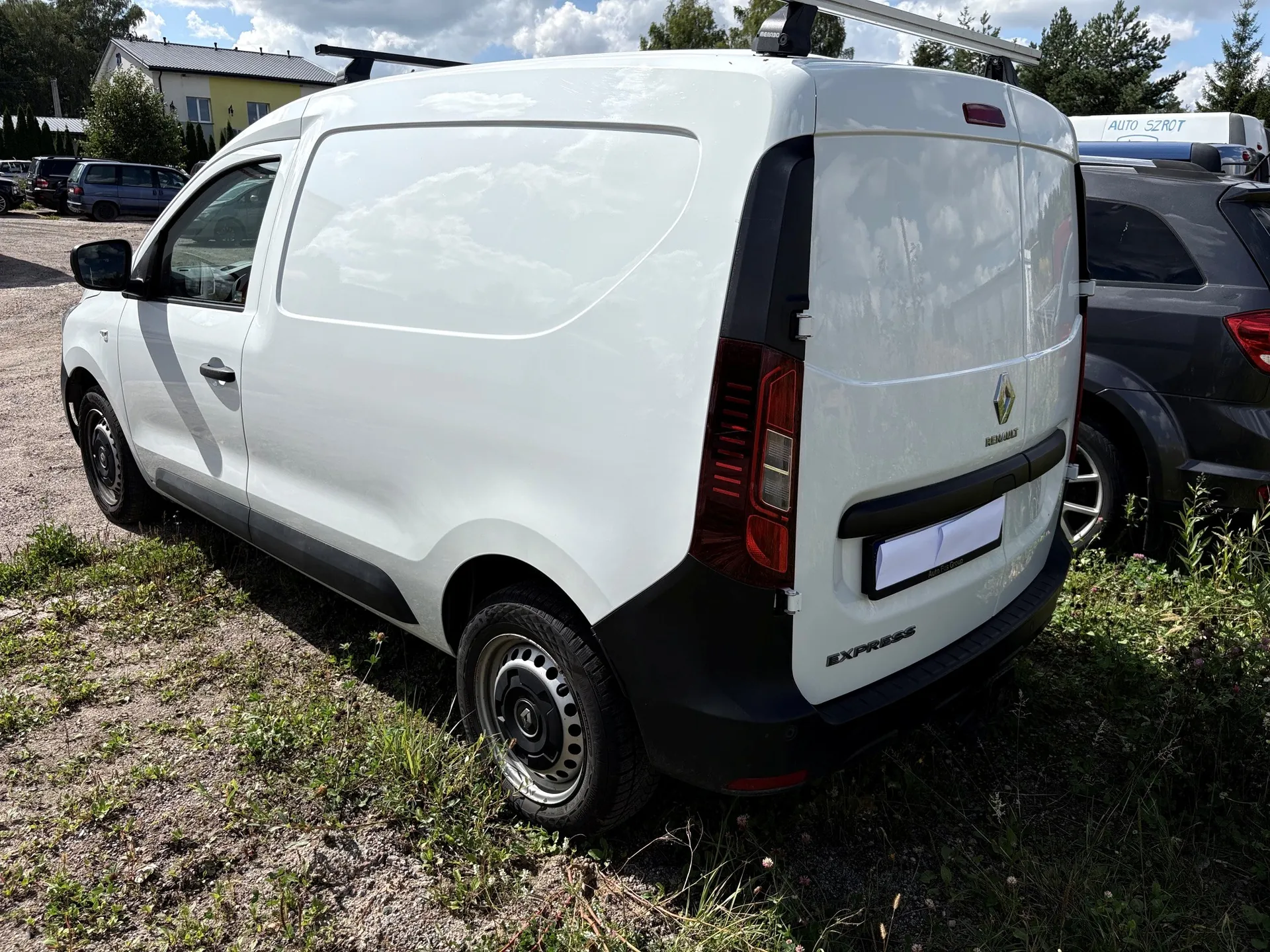 Biały samochód dostawczy marki Renault Express zaparkowany na trawiasto-żwirowym podłożu. Pojazd jest pokazany z tyłu i z boku. Na dachu zamontowany jest czarny bagażnik. W tle widoczne są inne samochody oraz budynek z napisem 'AUTO-SZROT'.