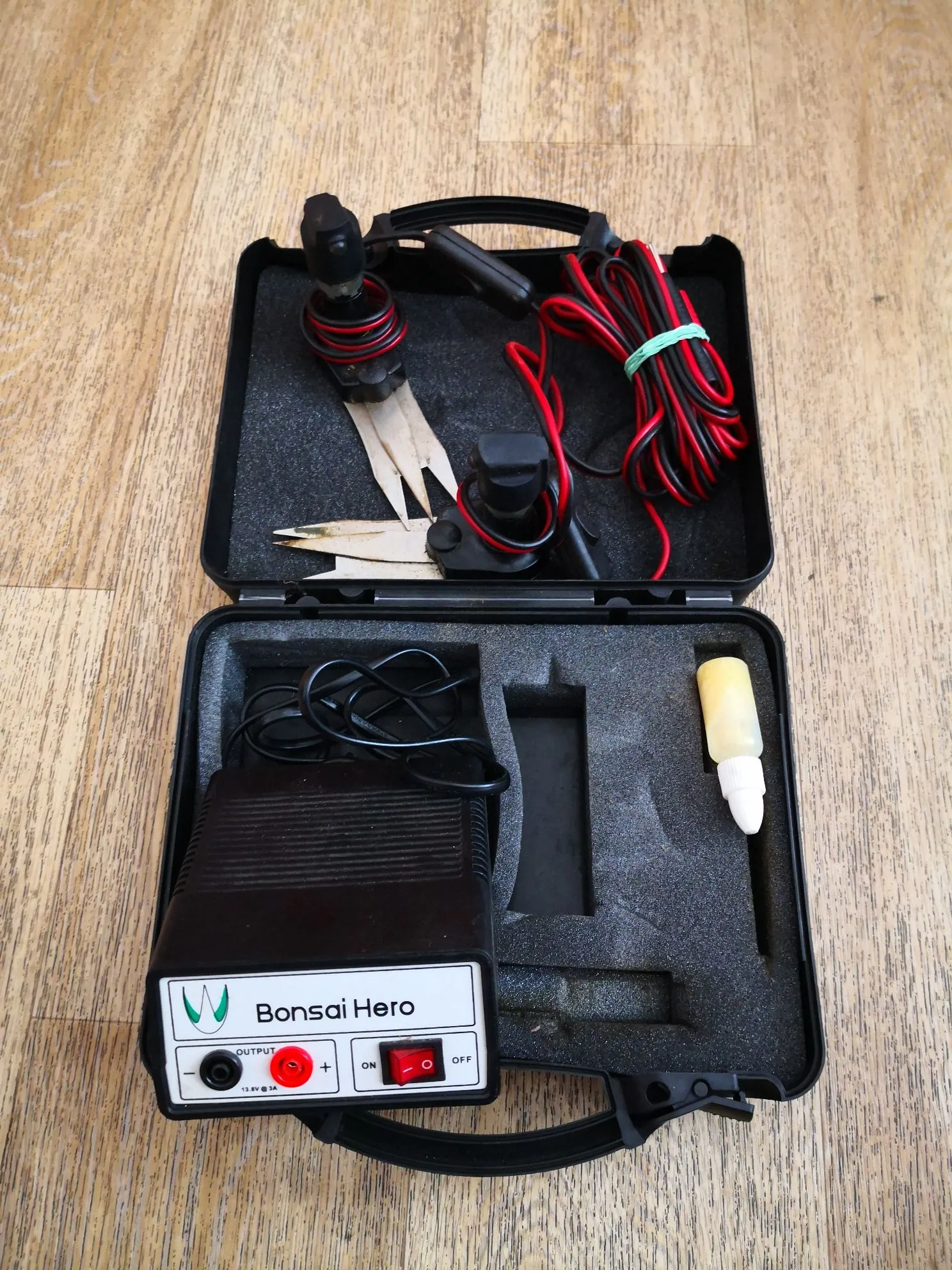 Otwarta czarna walizka z piankową wkładką, w której znajdują się narzędzia elektroniczne. W dolnej części znajduje się czarne urządzenie z białym panelem z napisem „Bonsai Hero”, czerwonym przyciskiem, przełącznikiem ON/OFF oraz gniazdami wyjściowymi. W górnej części walizki znajdują się czerwone i czarne przewody elektryczne, pęseta oraz inne drobne elementy techniczne. Po prawej stronie w piance umieszczony jest biały, stożkowaty element. Całość leży na drewnianej podłodze.