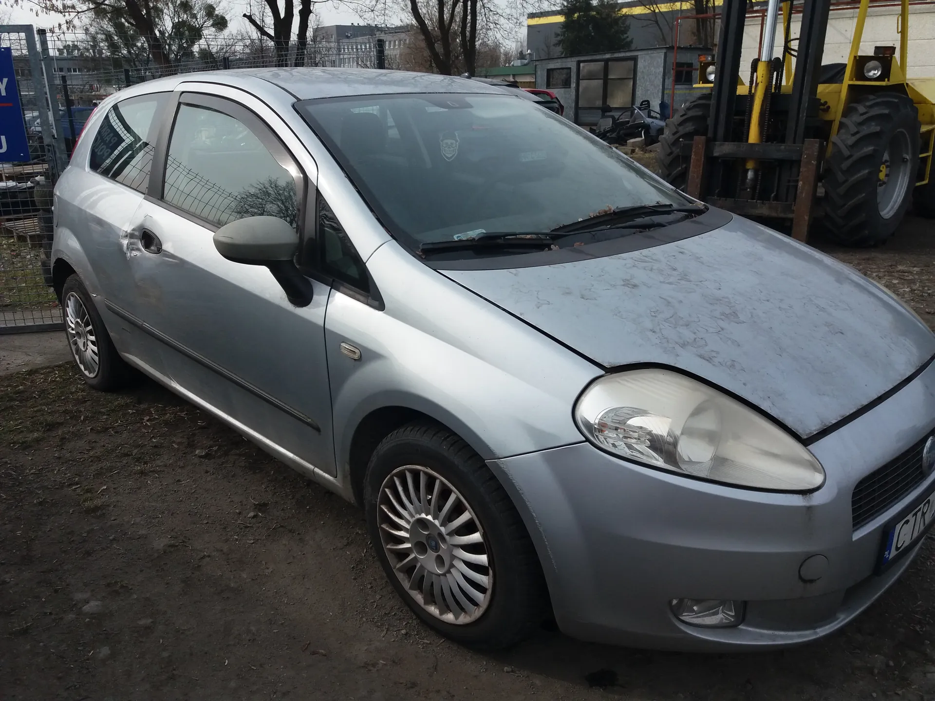 Srebrny samochód osobowy typu hatchback zaparkowany na brudnej, ziemistej powierzchni. Po lewej stronie widoczny jest metalowy płot z niebieskim znakiem zawierającym fragmenty liter "Y" i "J". Na przedniej szybie samochodu naklejka w kształcie czaszki. Na masce brud i ślady zanieczyszczeń. Po prawej stronie żółty wózek widłowy z dużymi oponami. W tle budynki, drzewa bez liści i inne pojazdy. Tablica rejestracyjna z częściowo widocznym numerem "CTR".