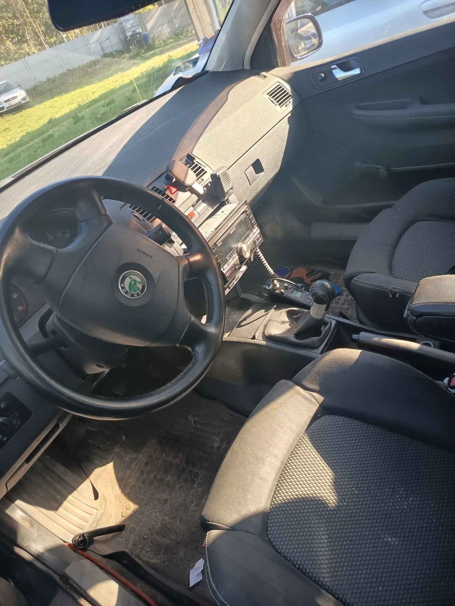 Wnętrze samochodu marki Skoda z perspektywy kierowcy. Widoczna jest czarna kierownica z logo marki, deska rozdzielcza z zamontowanym radiem, dźwignia zmiany biegów oraz dwa przednie fotele z ciemnej tkaniny. Przez przednią szybę widoczny jest fragment trawnika i budynku.