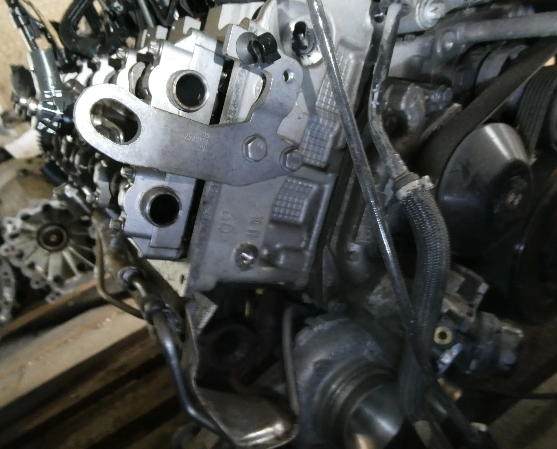Zbliżenie na metalowe części silnika spalinowego. Widoczne są aluminiowe odlewy, śruby, przewody oraz pas napędowy po prawej stronie. Na jednej z metalowych płytek znajduje się niewyraźny grawer.