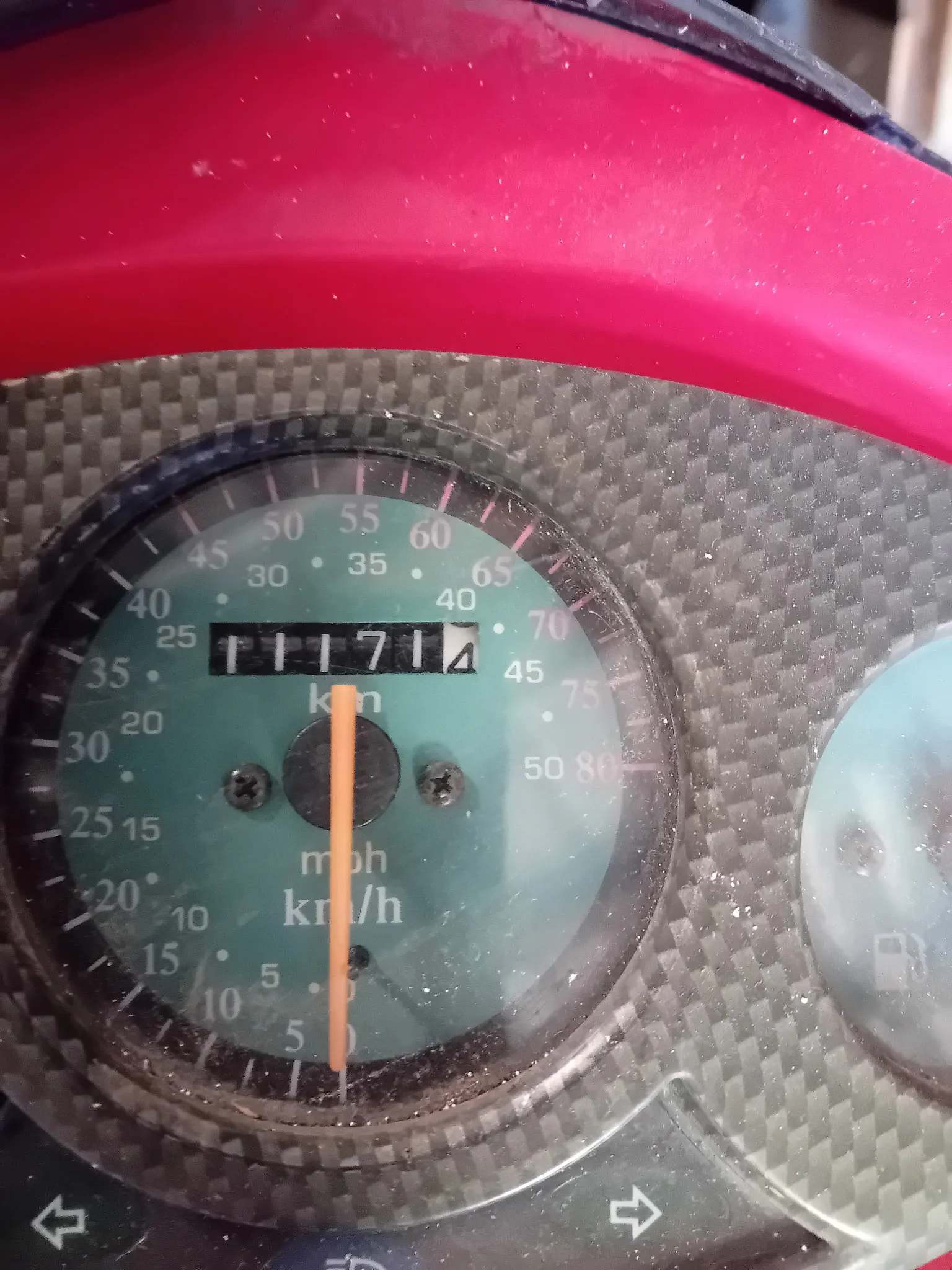 Zbliżenie na licznik prędkości w pojeździe. Tarcza licznika jest zielona z białymi cyframi wskazującymi prędkość w mph i km/h. Pomarańczowa wskazówka znajduje się w pozycji zero. W górnej części widoczny jest licznik przebiegu z wartością 11714. Tło wokół licznika ma strukturę włókna węglowego, a nad nim znajduje się czerwony element obudowy.