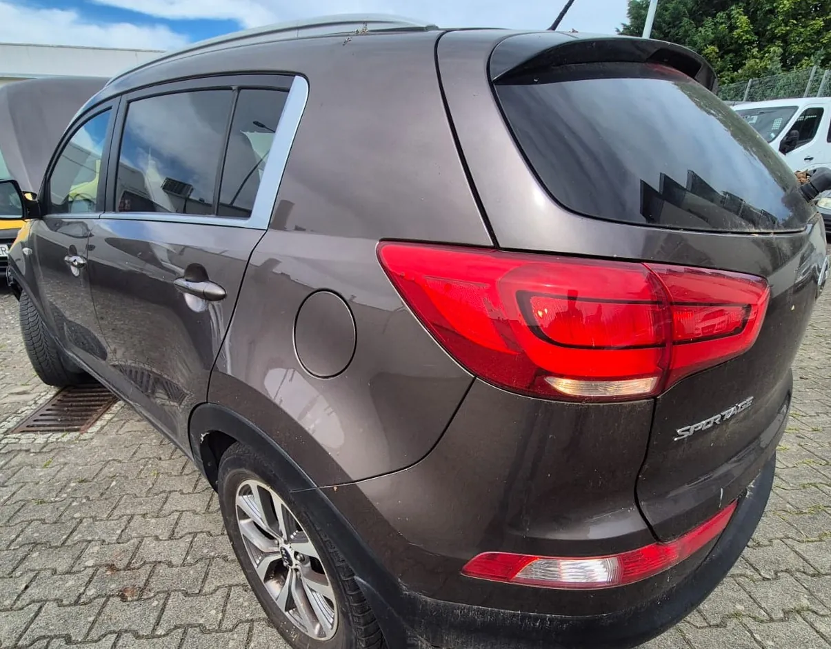 Tylna część brązowego samochodu marki Kia, model Sportage, zaparkowanego na kostce brukowej. Widoczny jest prawy tylny reflektor, klapka wlewu paliwa oraz napis 'SPORTAGE' na klapie bagażnika.