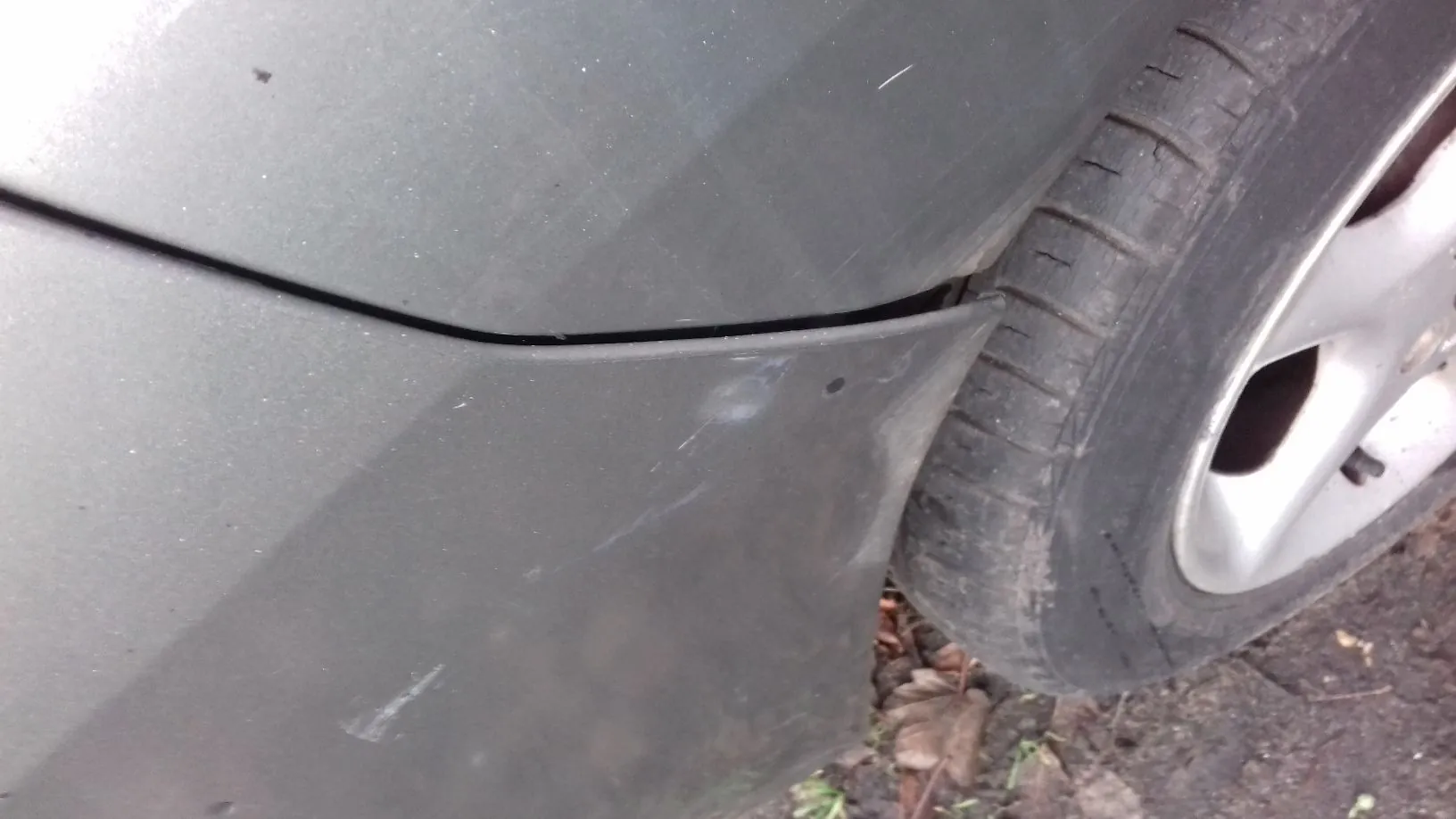 Zbliżenie na fragment ciemnoszarego nadwozia samochodu w okolicy przedniego lub tylnego koła. Widoczna jest szczelina między elementami karoserii oraz zarysowania i zabrudzenia na lakierze. Po prawej stronie znajduje się opona z felgą aluminiową, a pod samochodem widoczna jest ziemia z liśćmi.