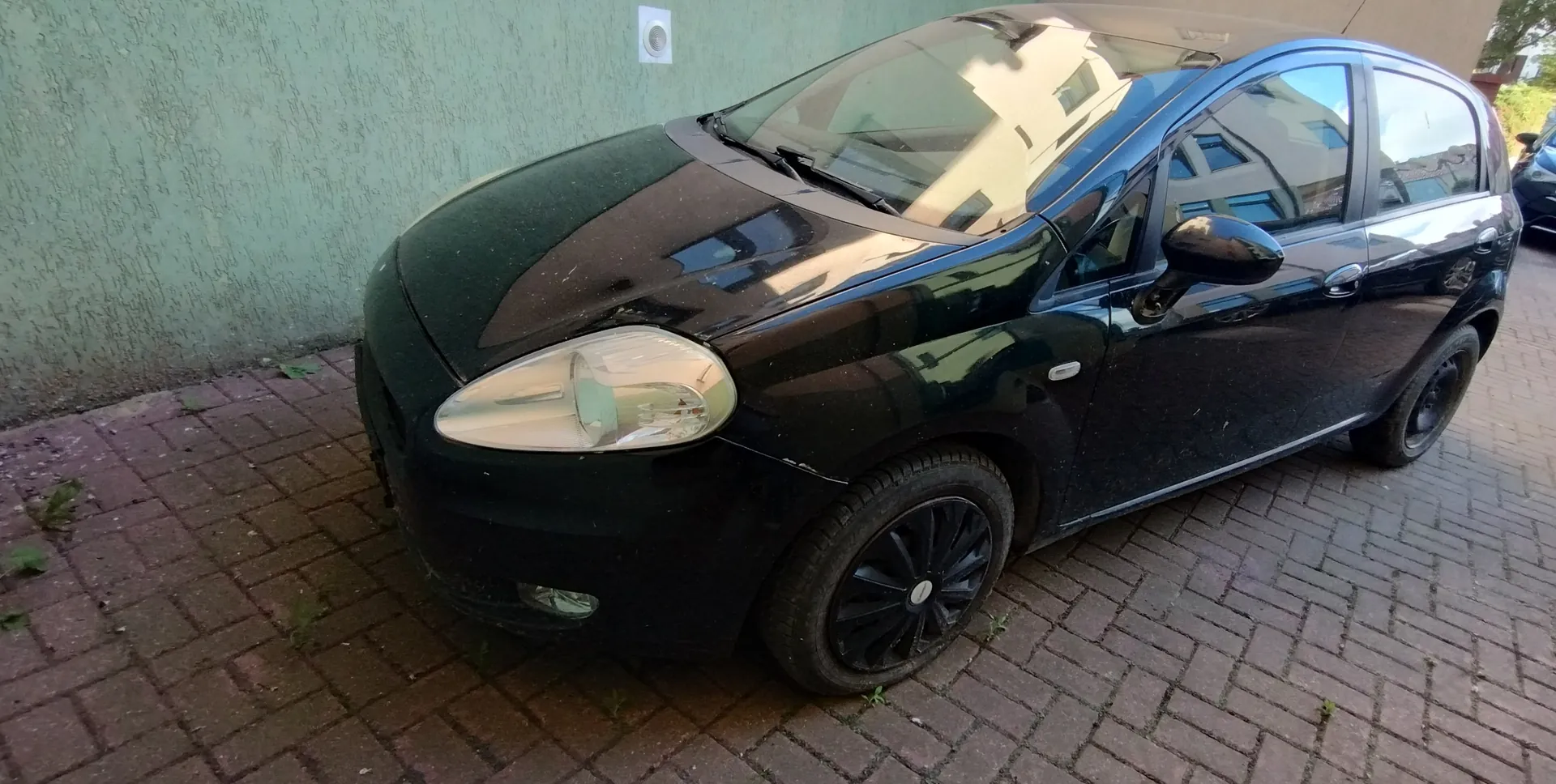 Czarny samochód osobowy typu hatchback zaparkowany na ceglanym chodniku przy zielonej, teksturowanej ścianie z białym kwadratowym elementem. Widoczne są reflektory, czarne felgi, lusterko boczne oraz odbicia budynków w szybach. Samochód znajduje się pod kątem do ściany, a na chodniku rosną pojedyncze rośliny.