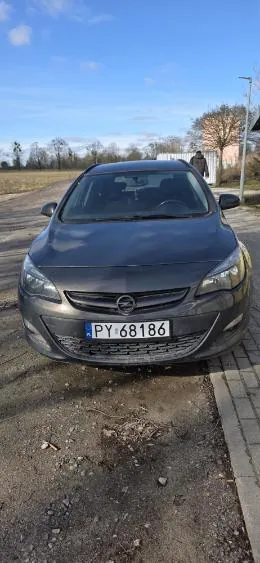 Czarny samochód marki Opel zaparkowany na drodze z żwiru obok chodnika z cegieł. Widoczna tablica rejestracyjna PY 68186. Tło: niebo z chmurami, drzewa i budynek. Horyzont poziomy, tekst na tablicy czytelny.