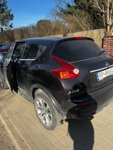 Czarny samochód Nissan Juke zaparkowany na brukowanej powierzchni. Drzwi kierowcy są otwarte. Widoczna tablica rejestracyjna RP 4964 oraz napis JUKE na tylnej części pojazdu. W tle drzewa, ceglana ściana i niebo. Cień osoby fotografującej widoczny na ziemi.